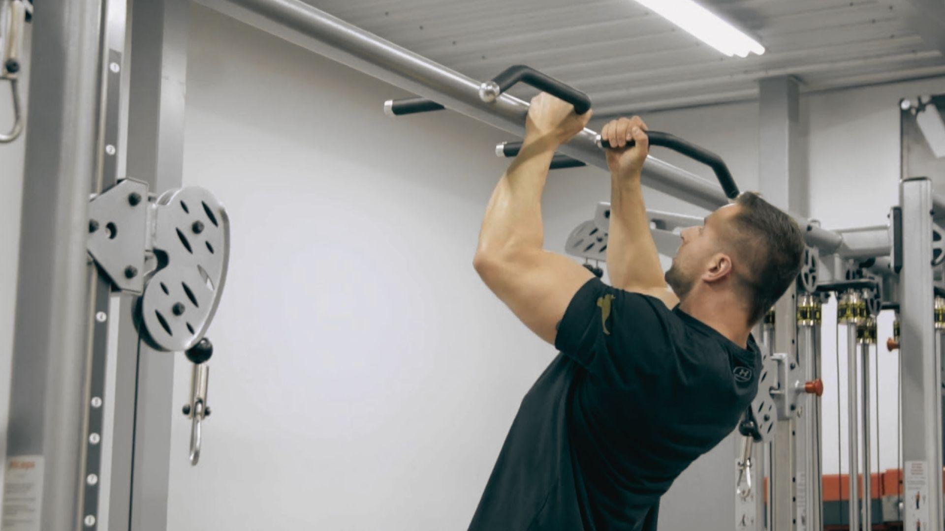 Man in black shirt doing neutral grip pull-up on cable machine pull-up bar, gym interior, fluorescent lights, profile
