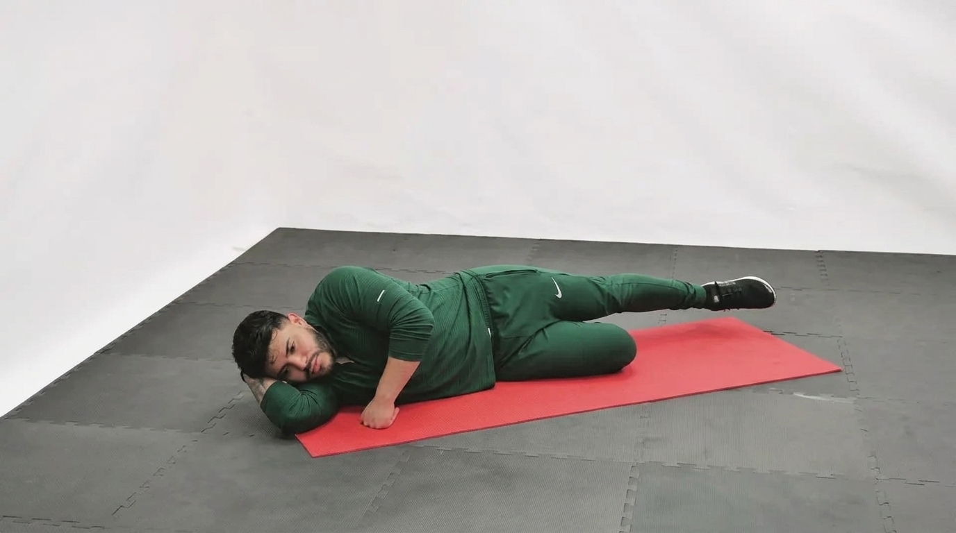 Man in green tracksuit lying on his side on a red mat performing a side-lying leg raise.