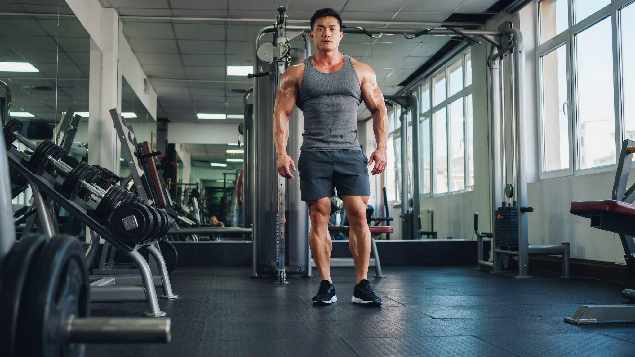 Muscular athlete in gray tank top walks in gym, broad shoulders and arms contrast with thinner legs.
