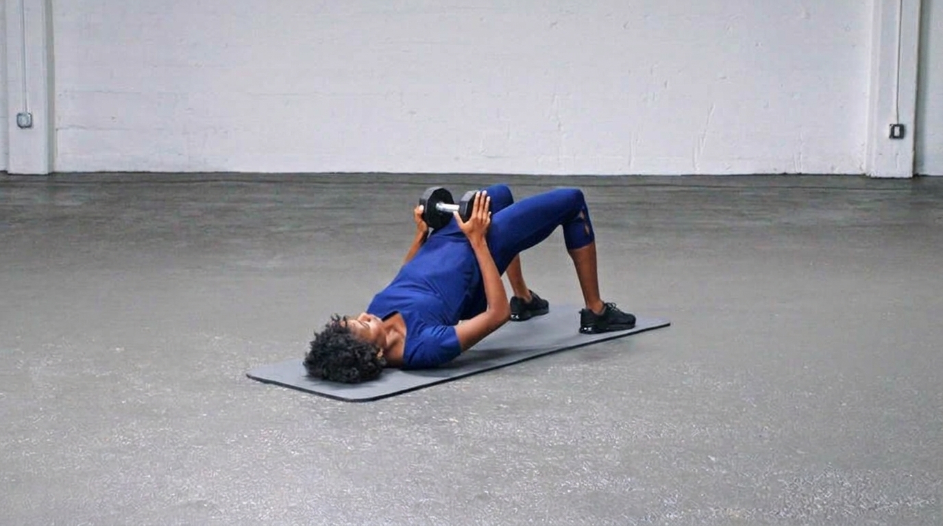 Woman in blue activewear does a dumbbell glute bridge on a mat, holding a dumbbell on her hips.