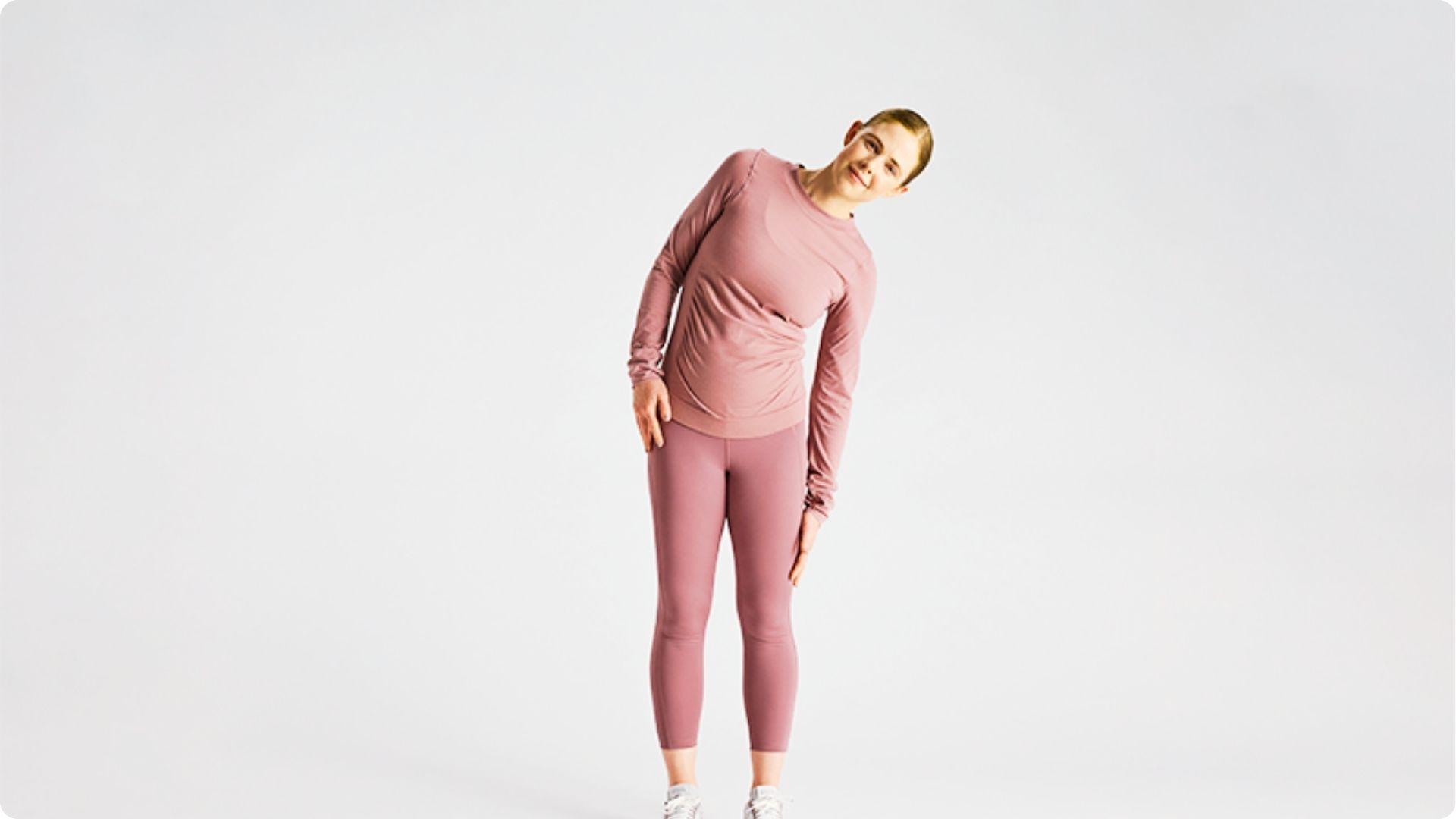 Woman in pink activewear stands against a white backdrop, bending sideways to perform a standing side reach.