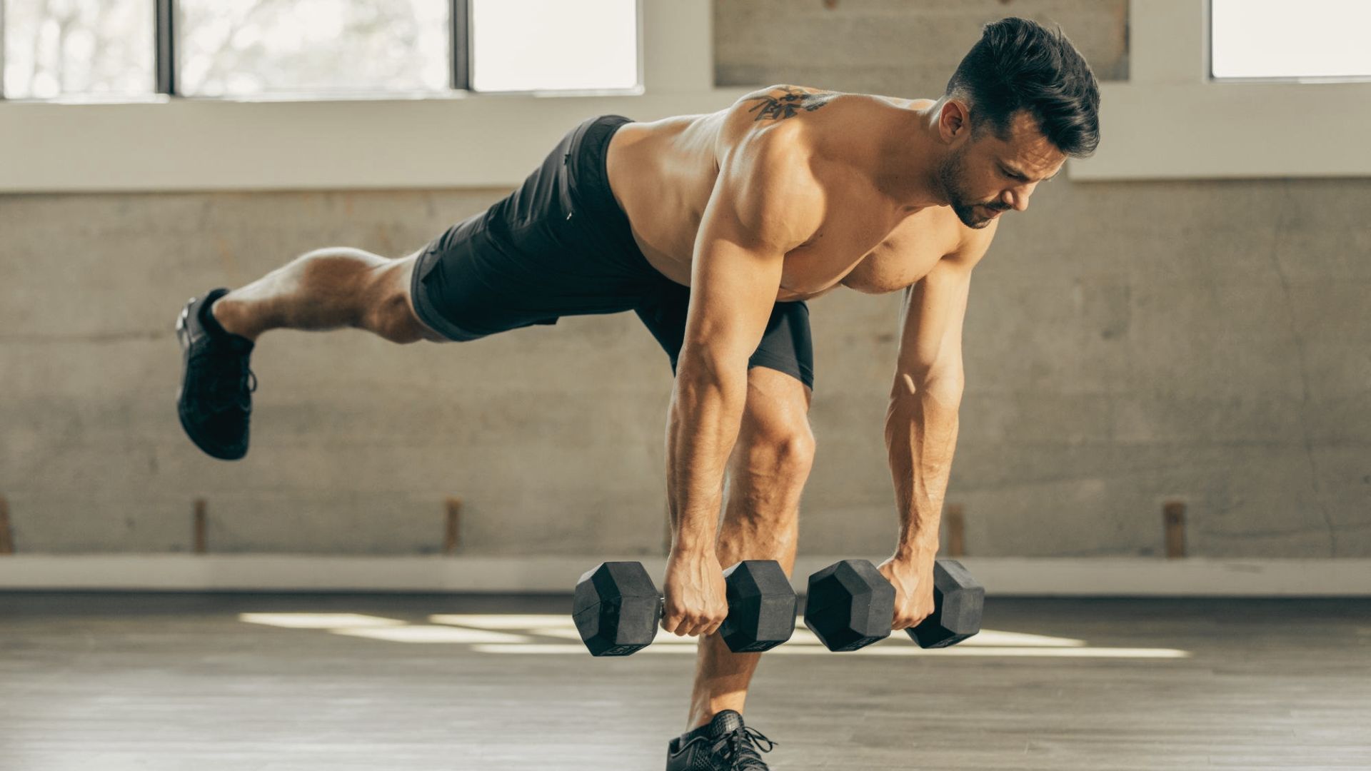 A shirtless man balances on one leg while hinging at the hips with dumbbells in both hands.