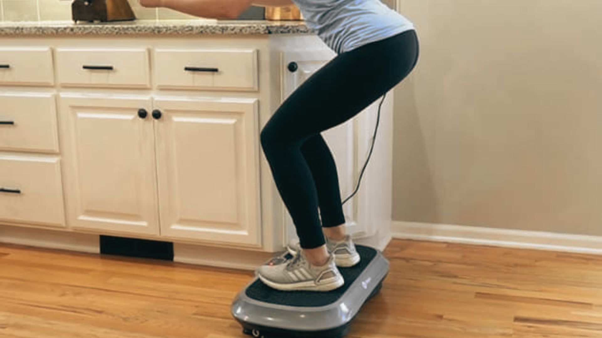 Close-up of a woman in black leggings and sneakers performing a squat on a compact vibration plate in a kitchen.