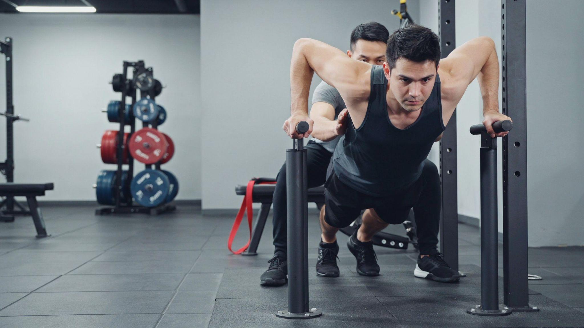 A man performing chest dips on parallel bars in a gym while a spotter assists him from behind.