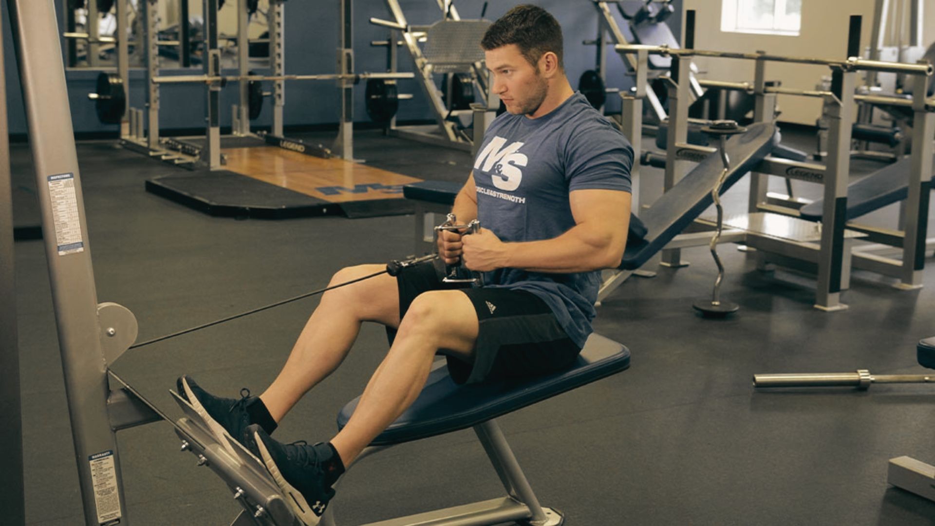 Athlete wearing a blue “Muscle & Strength” shirt doing a seated cable row with a V-bar handle.