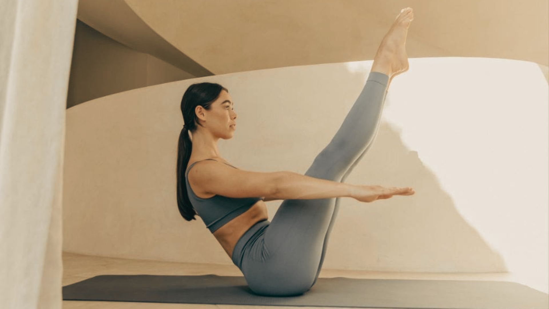 Woman in grey activewear holds a Pilates Teaser on a mat, balancing on her sit bones with legs extended and arms reaching forward.