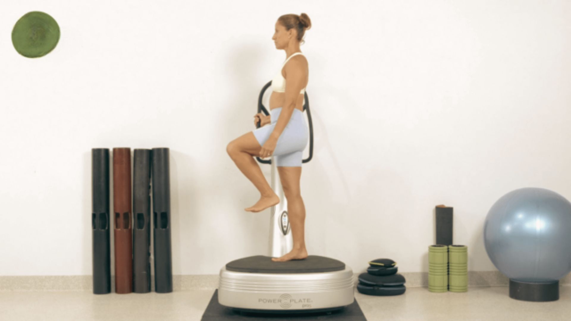 Woman in a white sports bra and light blue shorts balances on one leg on a Power Plate machine.