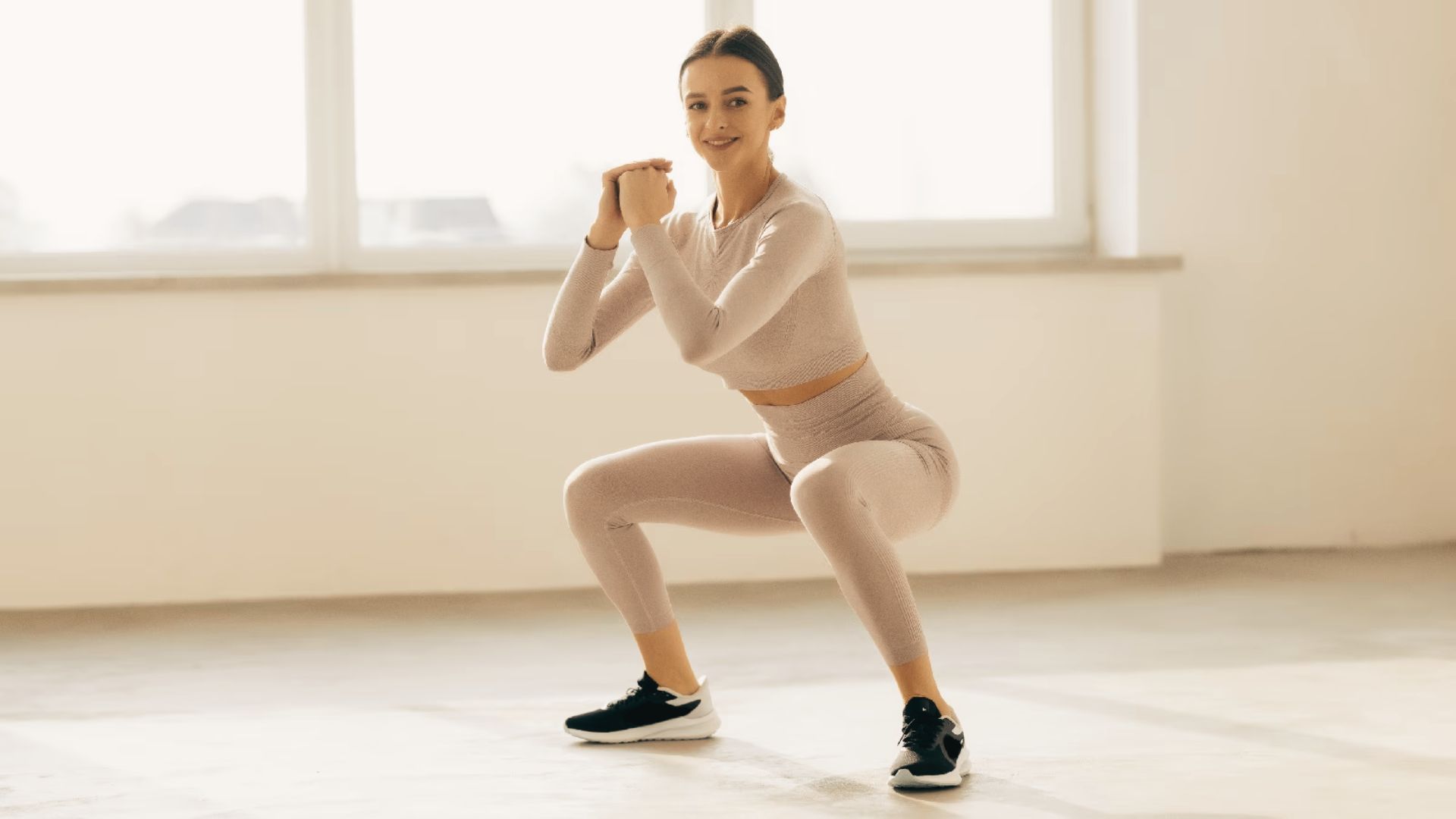 Woman in light workout set performing a bodyweight squat with hands clasped near her chest.