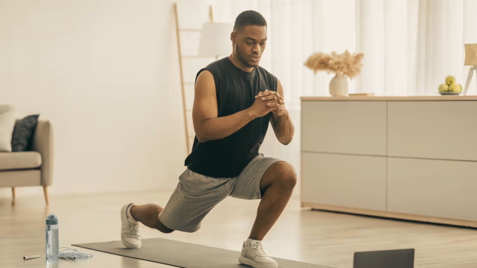 Man in black tank top and gray shorts doing a reverse lunge on a mat in a bright living room.