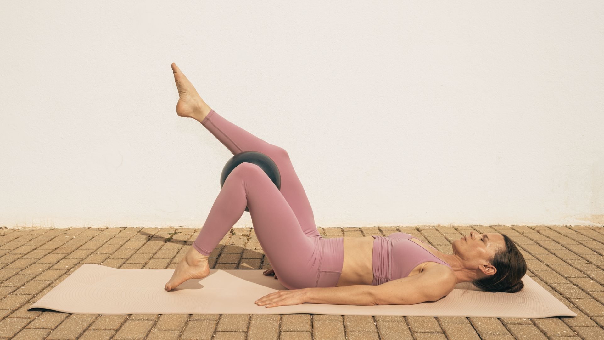 Woman in mauve activewear on a mat using a small Pilates ball between her knees.