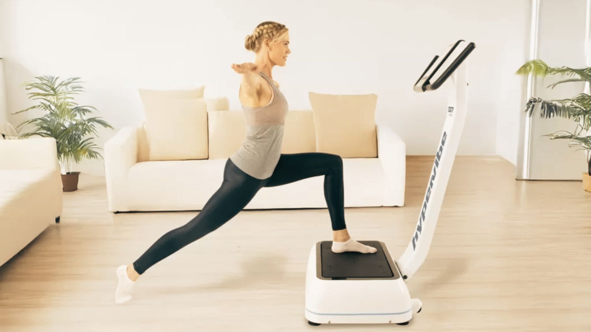 Woman in grey tank top and black leggings does a lunge with one foot on a Hypervibe vibration plate in a bright living room.