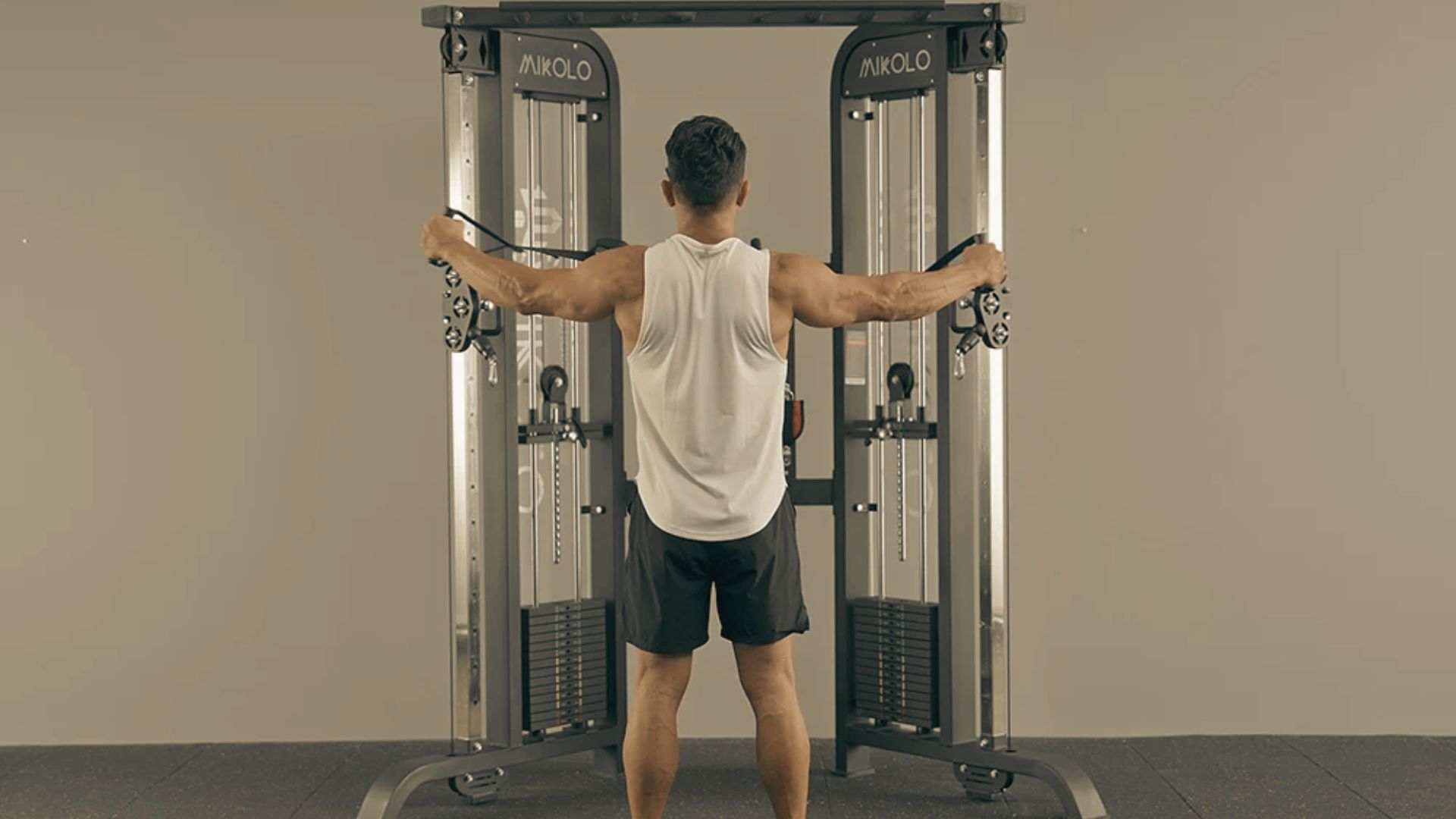 A man in a white tank top stands at a cable crossover machine performing a reverse fly.