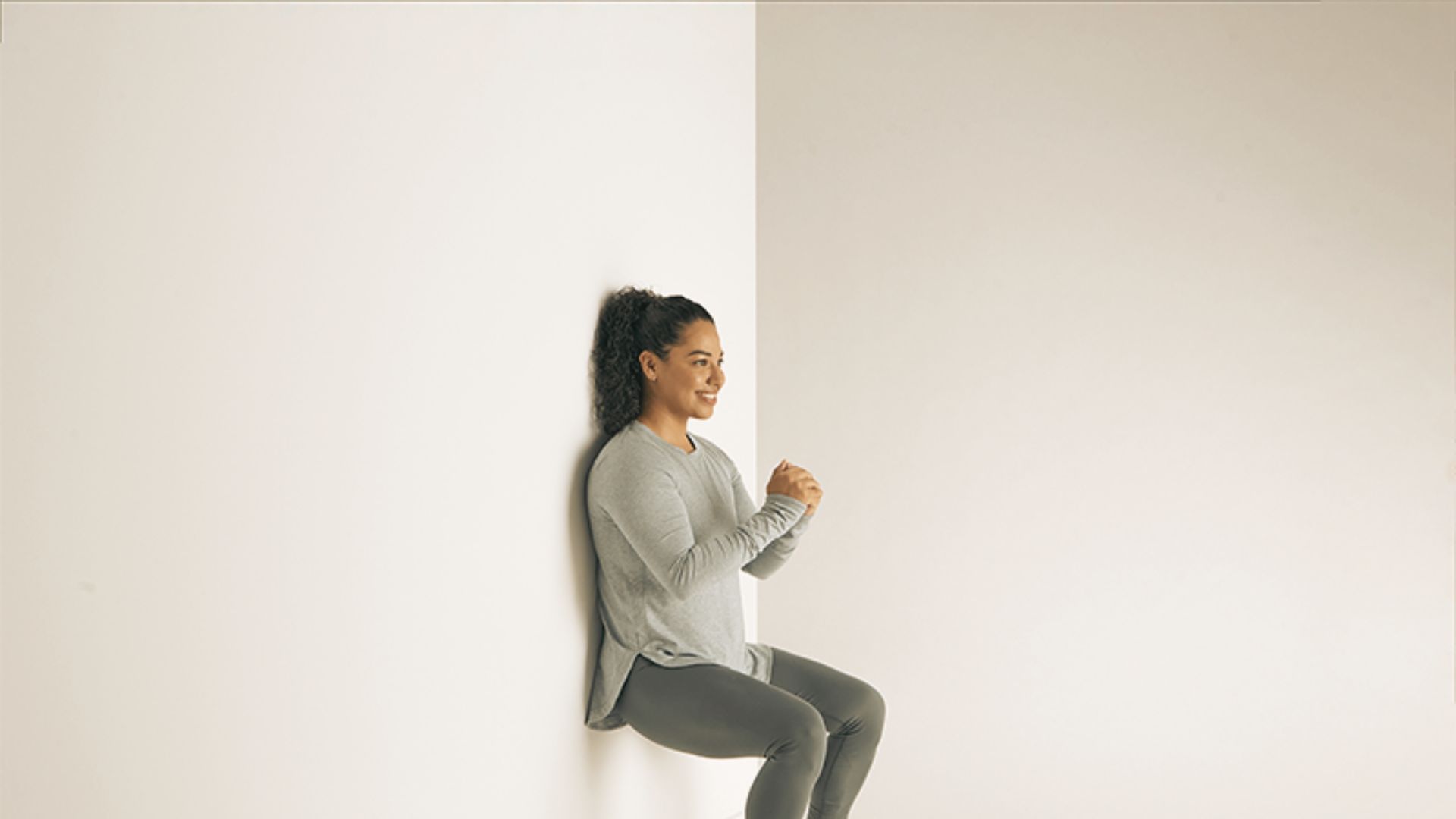 Woman in grey long-sleeve top and leggings holds a wall sit position against a white wall.