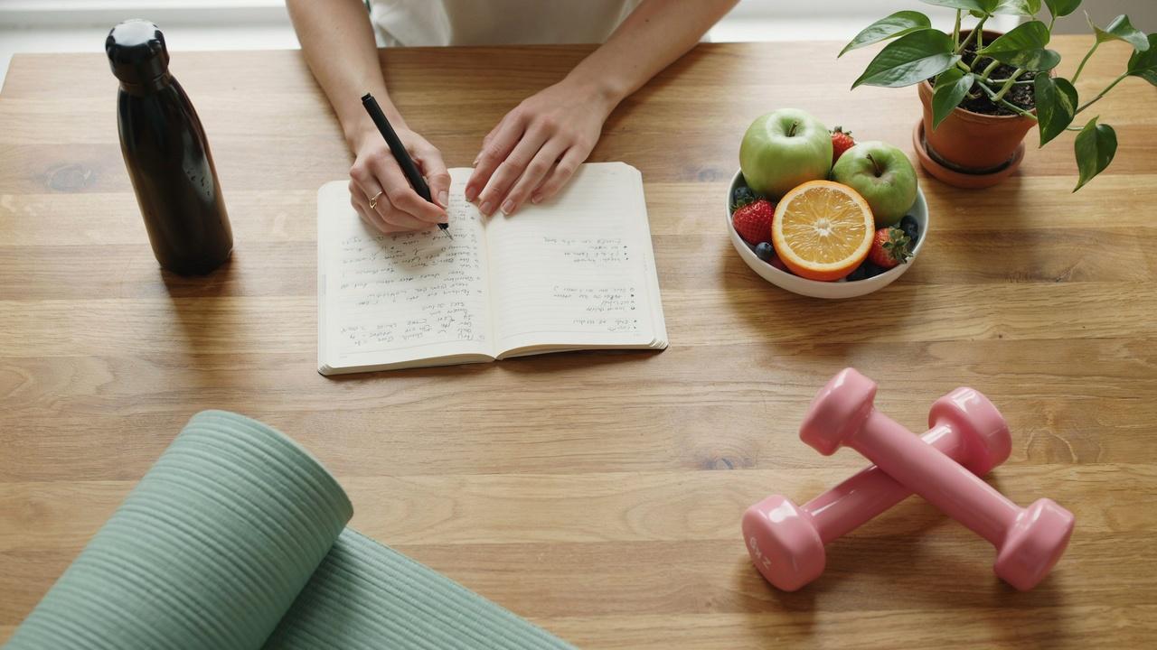 A woman writing fitness goals in a notebook with dumbbells, fruits, yoga mat and water bottle nearby.