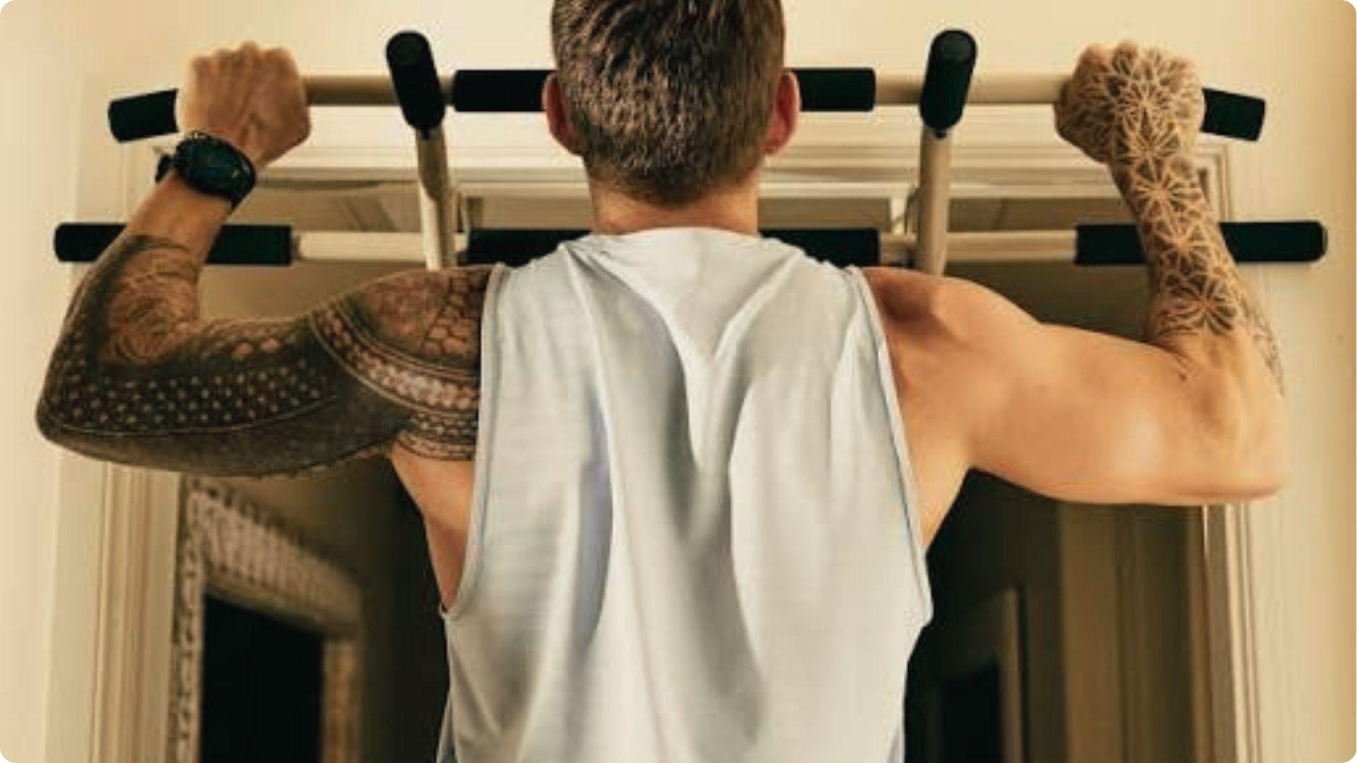 Tattooed man in white tank using doorway pull-up bar, targeting lats and rear delts