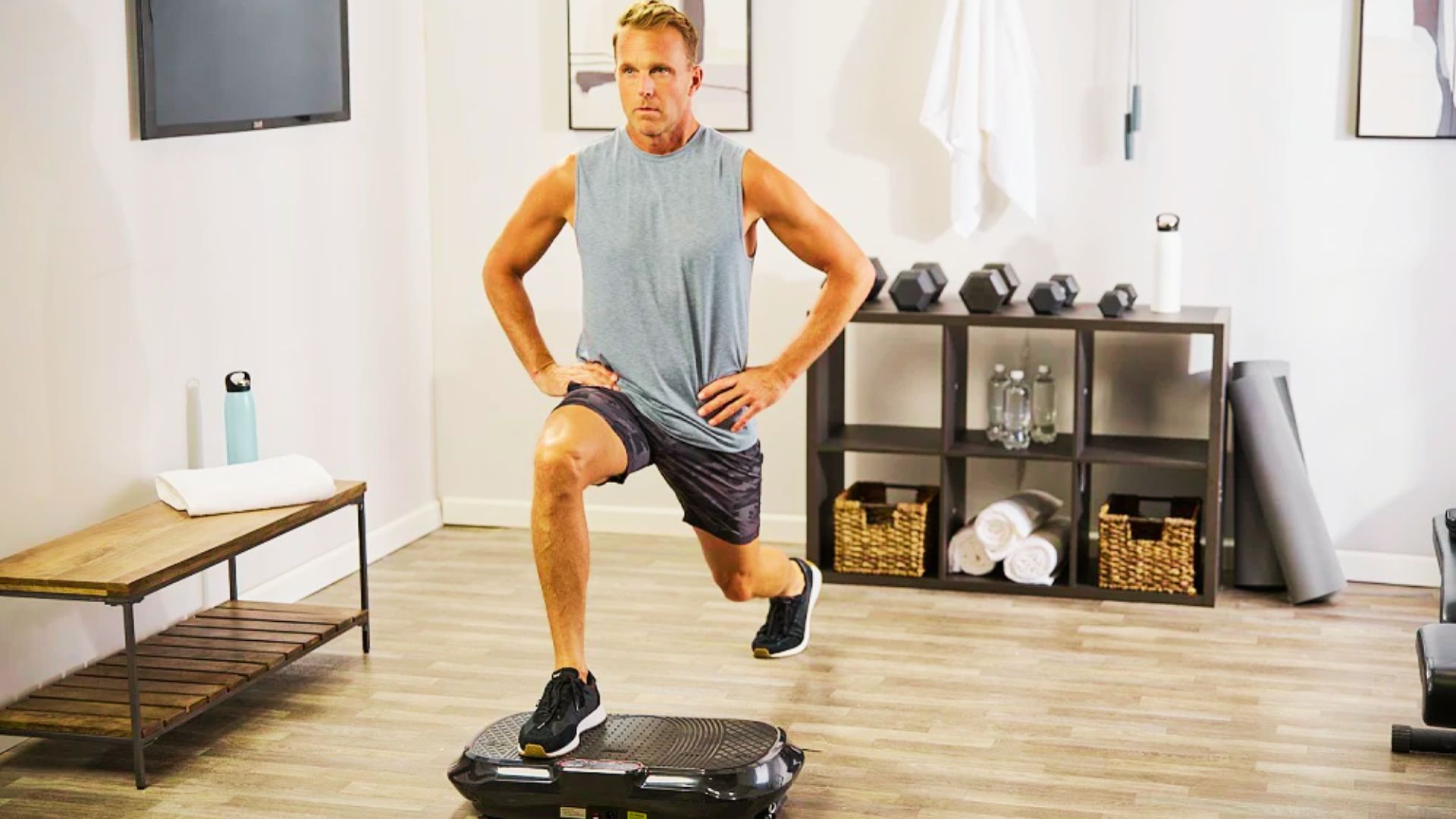 A man in a gray sleeveless shirt and camo shorts performs a static lunge with his right foot on a black vibration plate in a home gym.