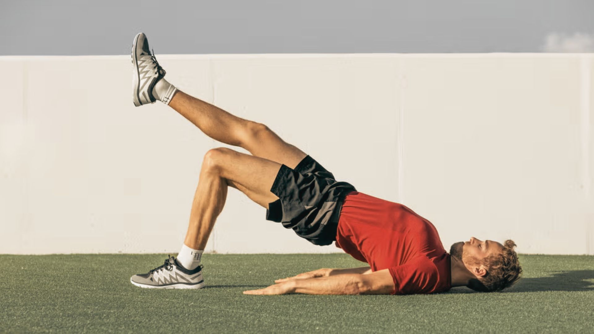 A man lies on turf raising his hips into a single-leg glute bridge with one leg extended upward.
