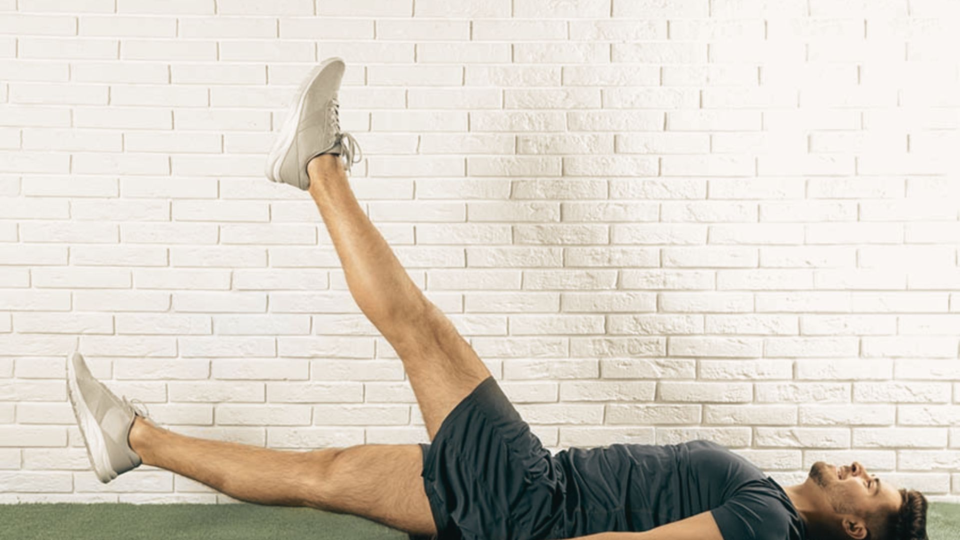 Man in black athletic wear and white sneakers lying on a mat doing Pilates scissor kicks.