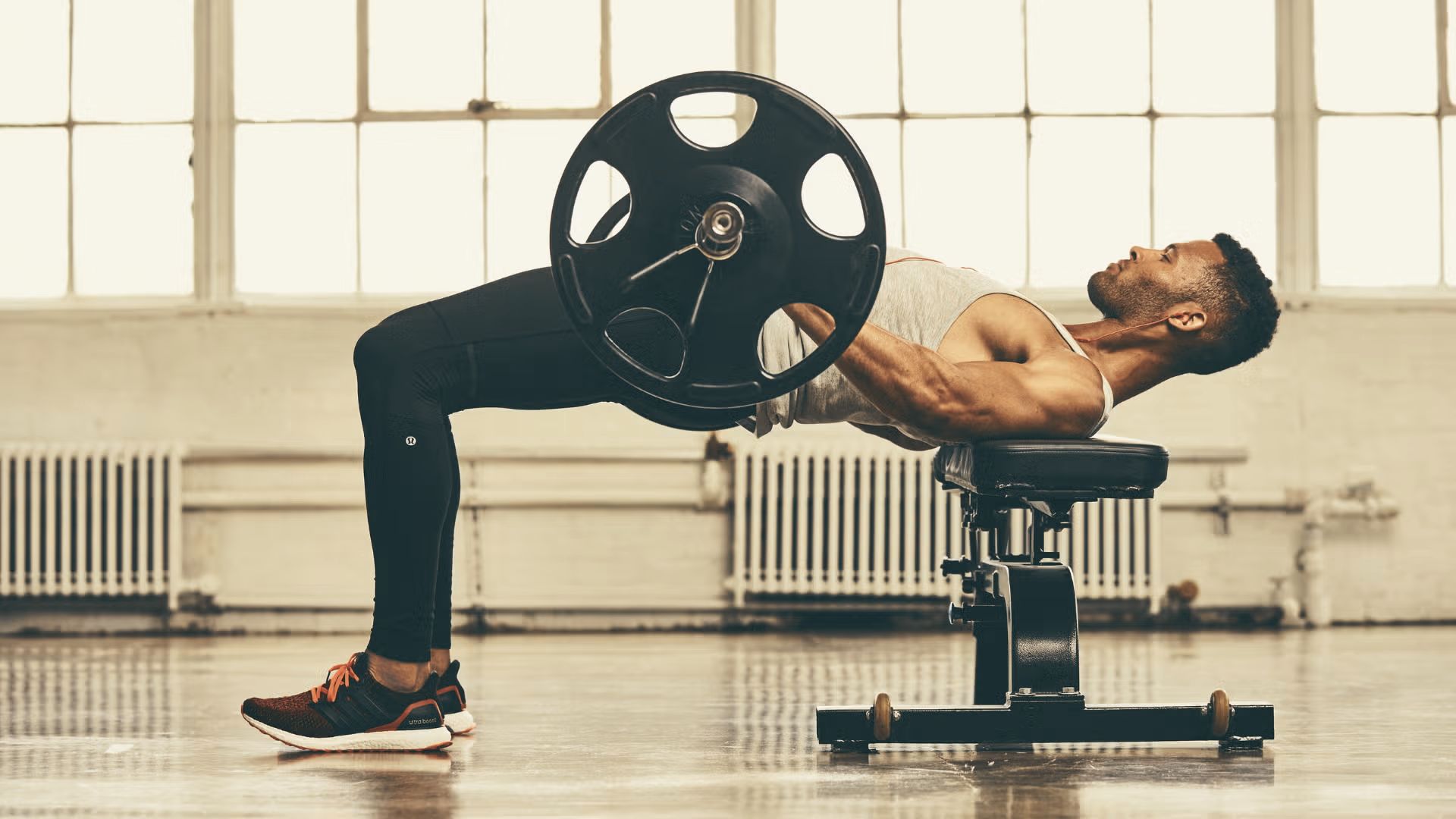 Man lying on a bench doing a barbell hip thrust in a bright gym studio.