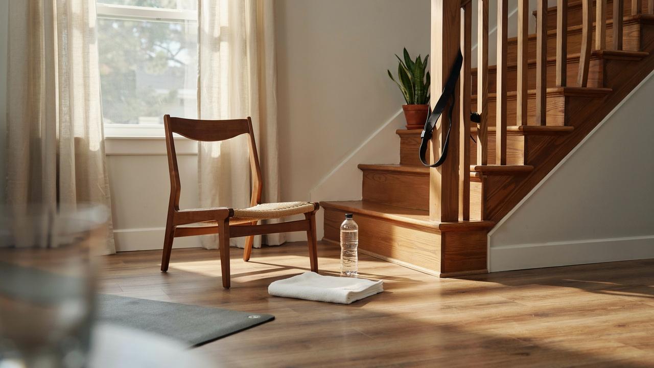 A home workout setup with a water bottle, chair, towel, resistance band looped around a stair railing, and staircase as fitness tools.