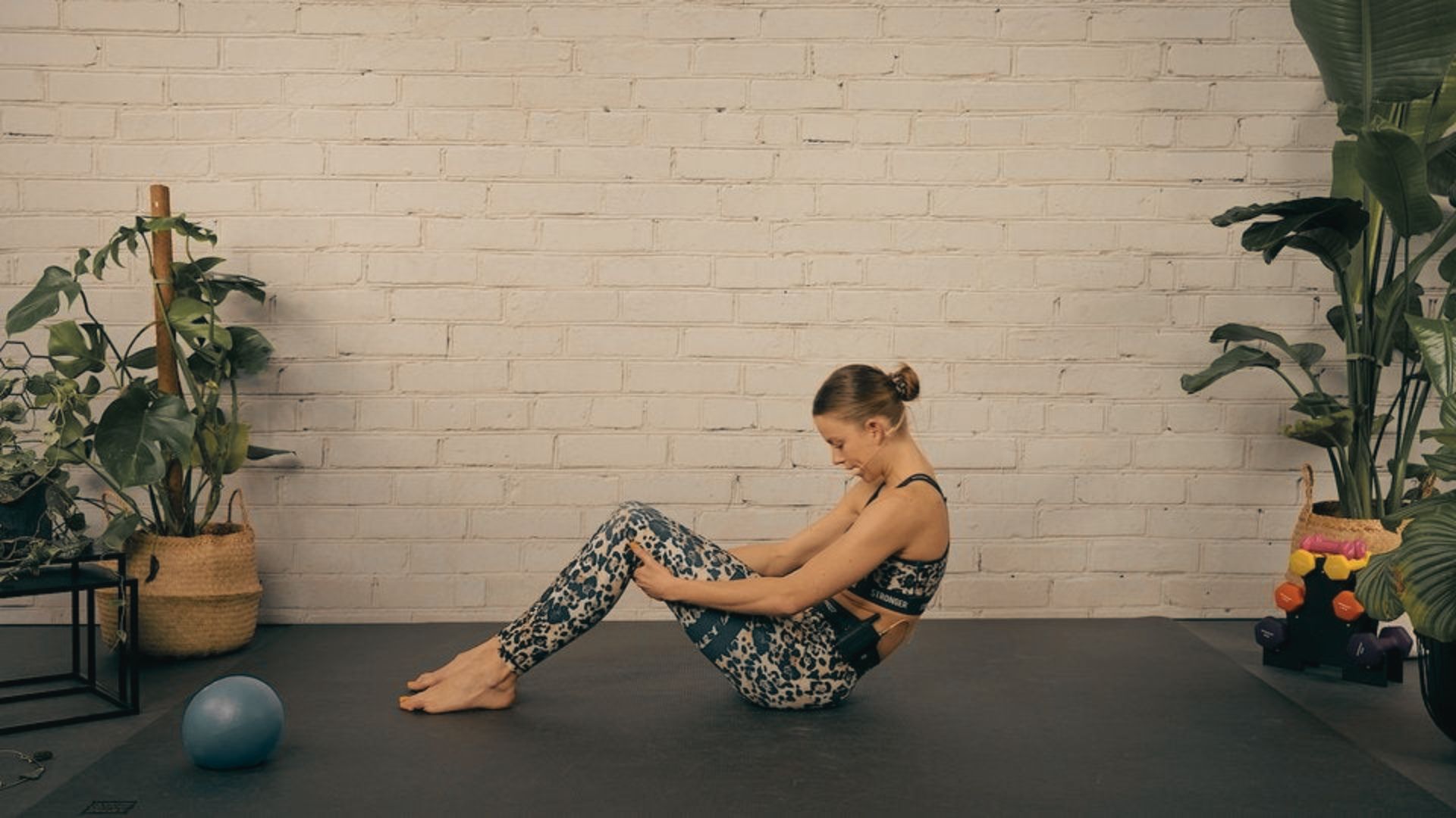 Woman sits on a mat in a bright studio with plants, leaning back slightly with bent knees and hands resting on her thighs.