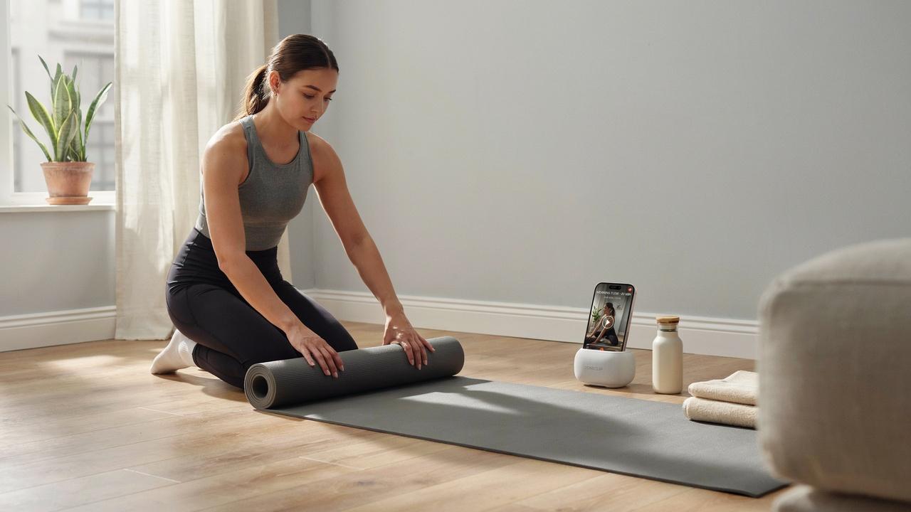 A person rolling out a yoga mat on the floor indoors, with a smartphone propped up next to a small bluetooth speaker.