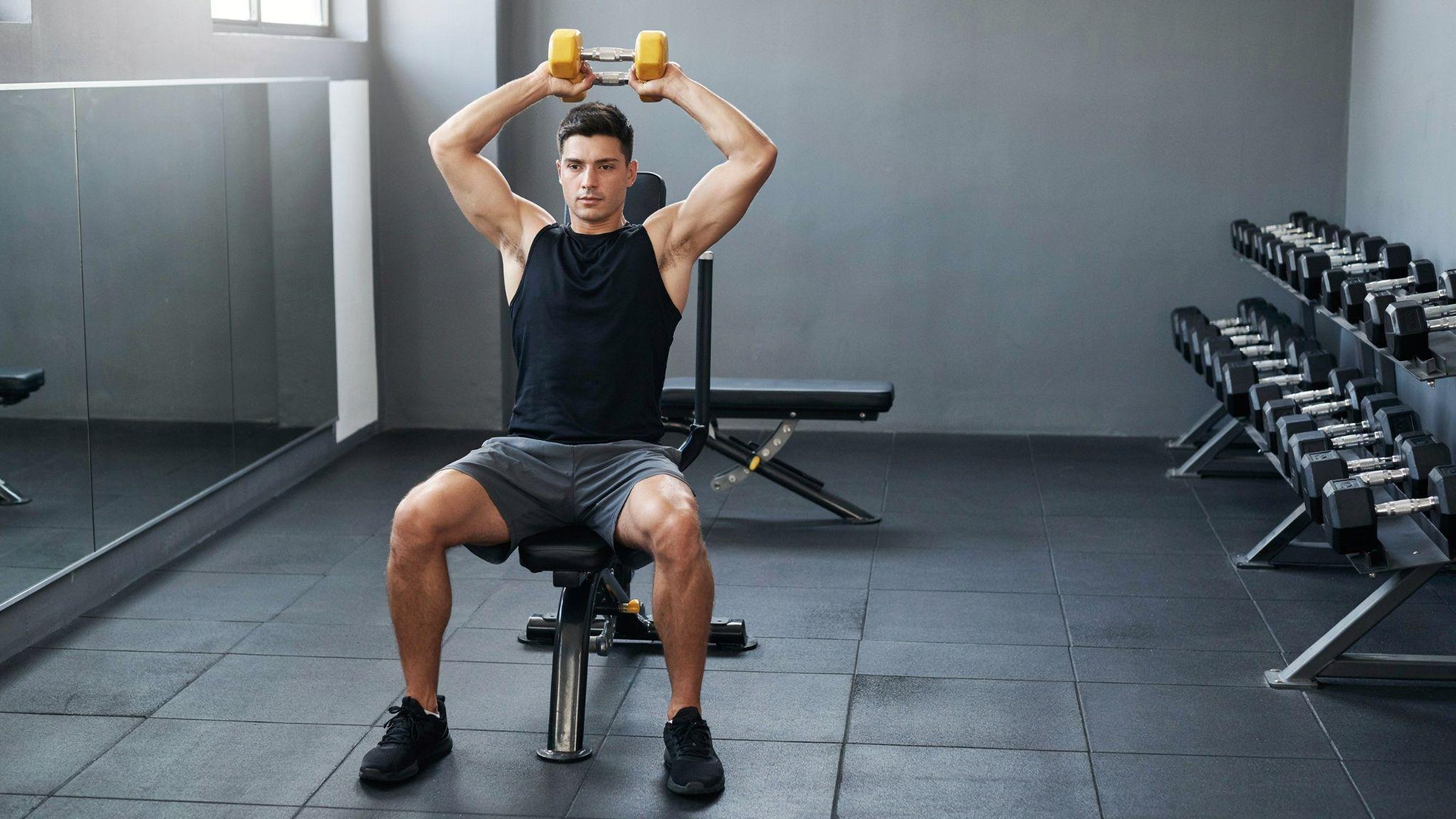 A man performing an overhead triceps extension with a dumbbell inside a gym.