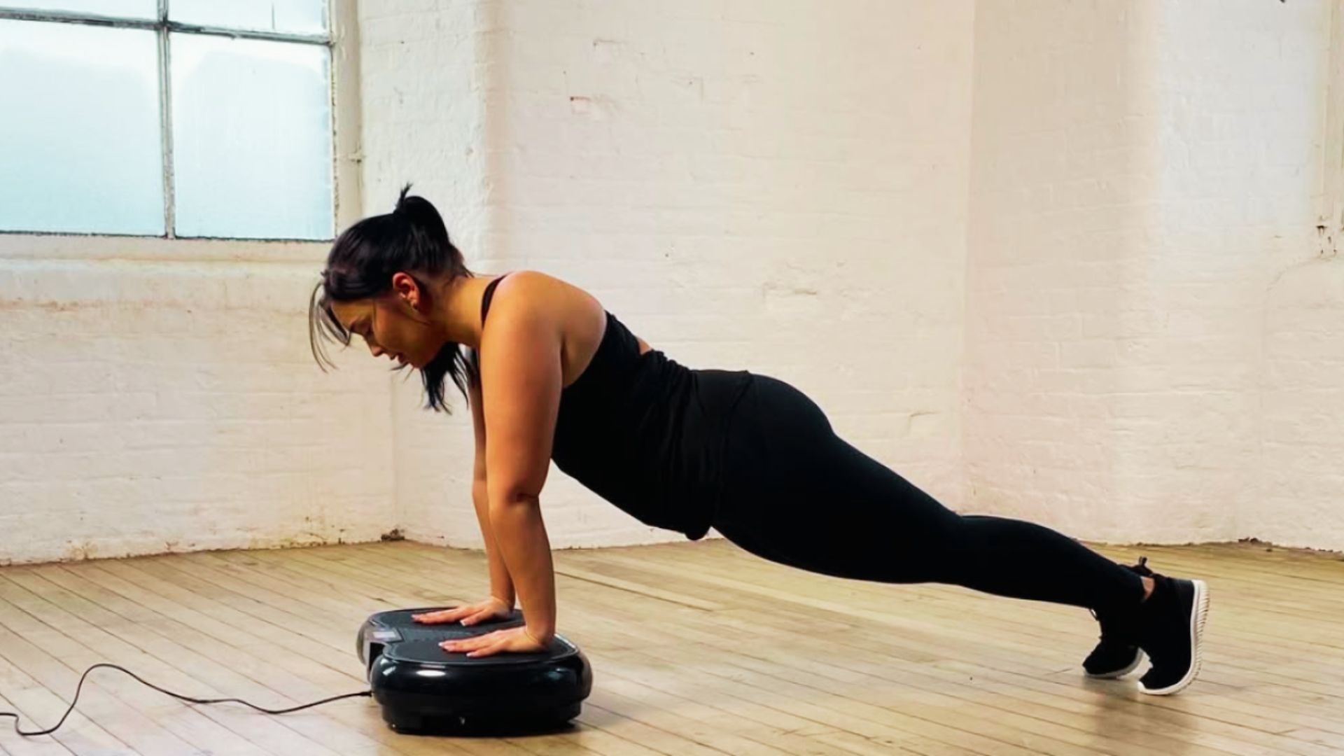 A woman in black workout attire holds a high plank with both hands placed on a black vibration plate in a bright studio with wood floors.