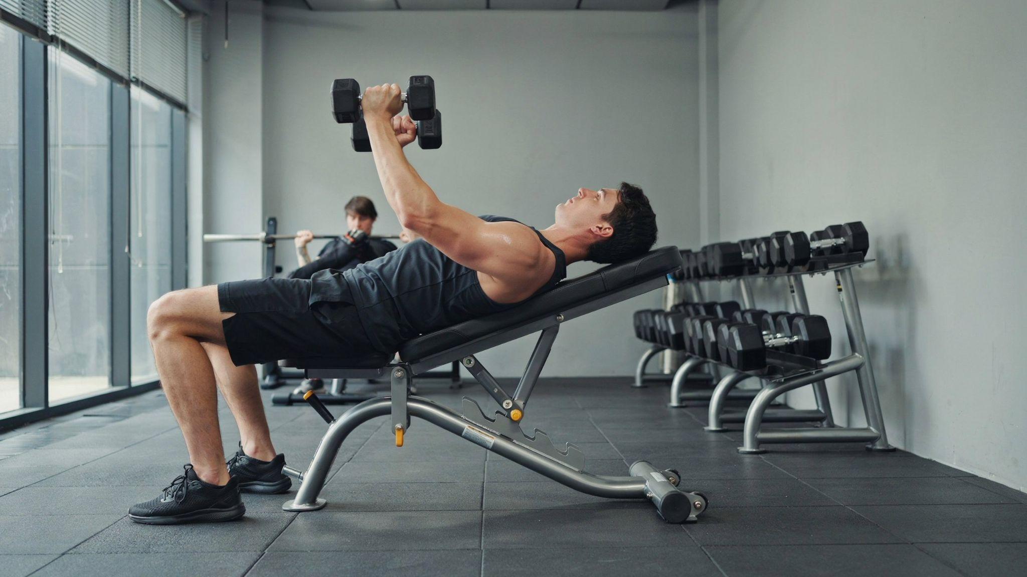 A man performing an incline dumbbell press on a weight bench inside a gym.
