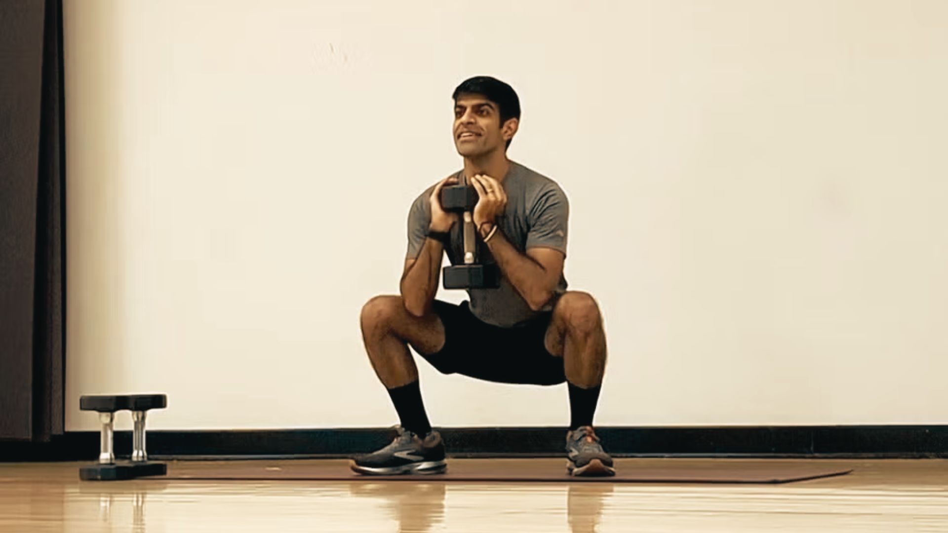 A man holds a dumbbell at his chest while performing a goblet squat on a mat indoors.