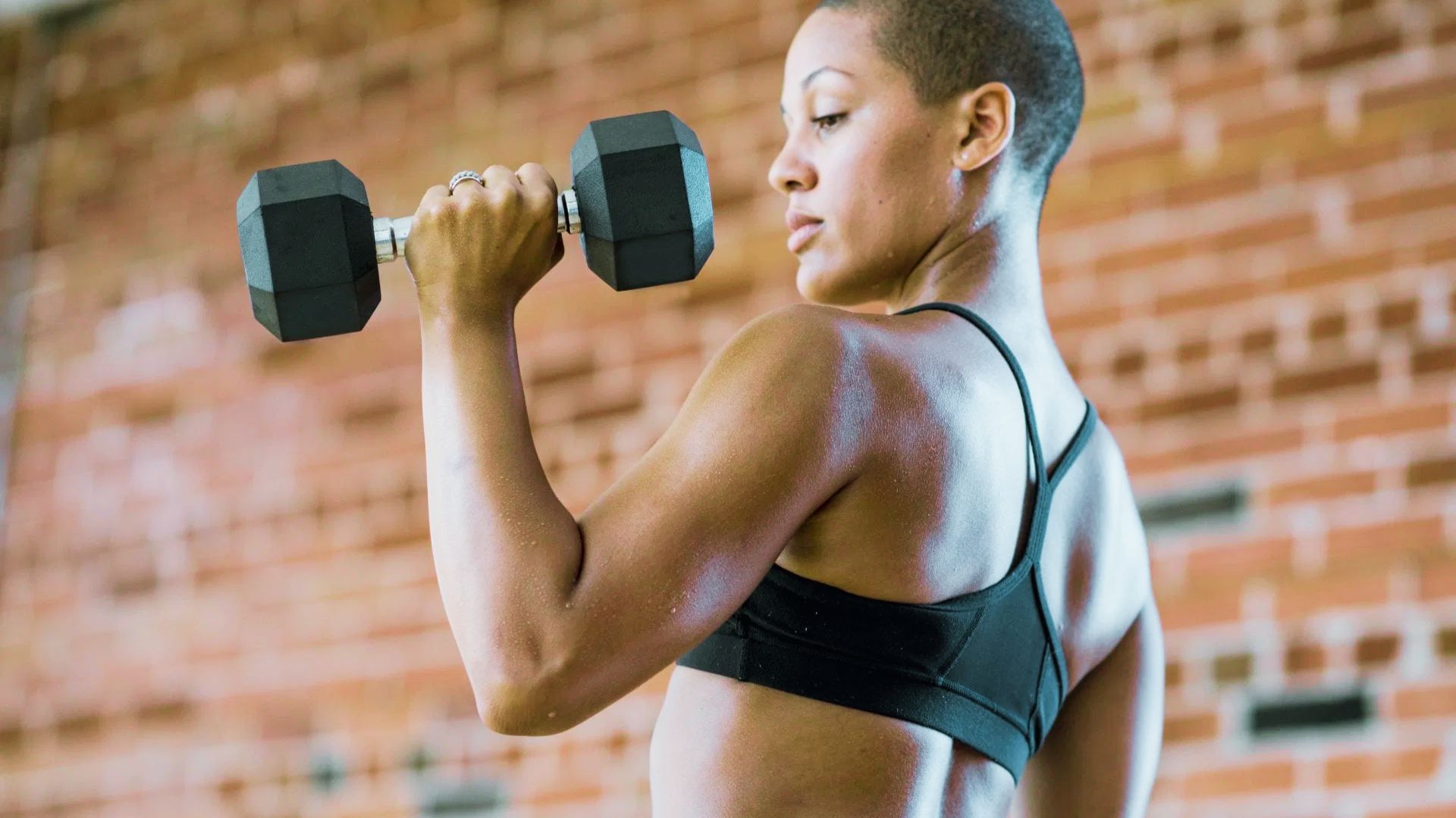 A woman with short hair lifts a black dumbbell in a bicep curl against a brick wall.