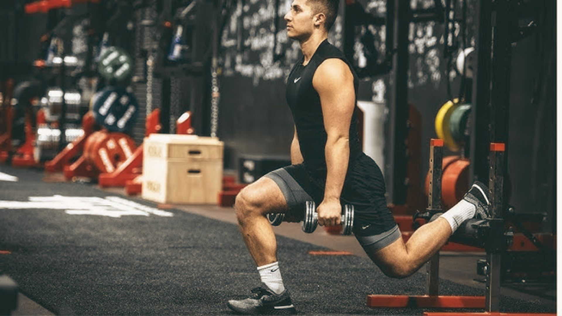 A man in a gym performs a Bulgarian split squat with dumbbells in each hand.