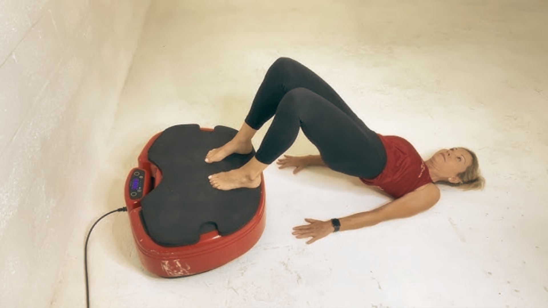 Woman in a red tank top and black leggings lies on the floor doing a glute bridge with feet on a red vibration plate.