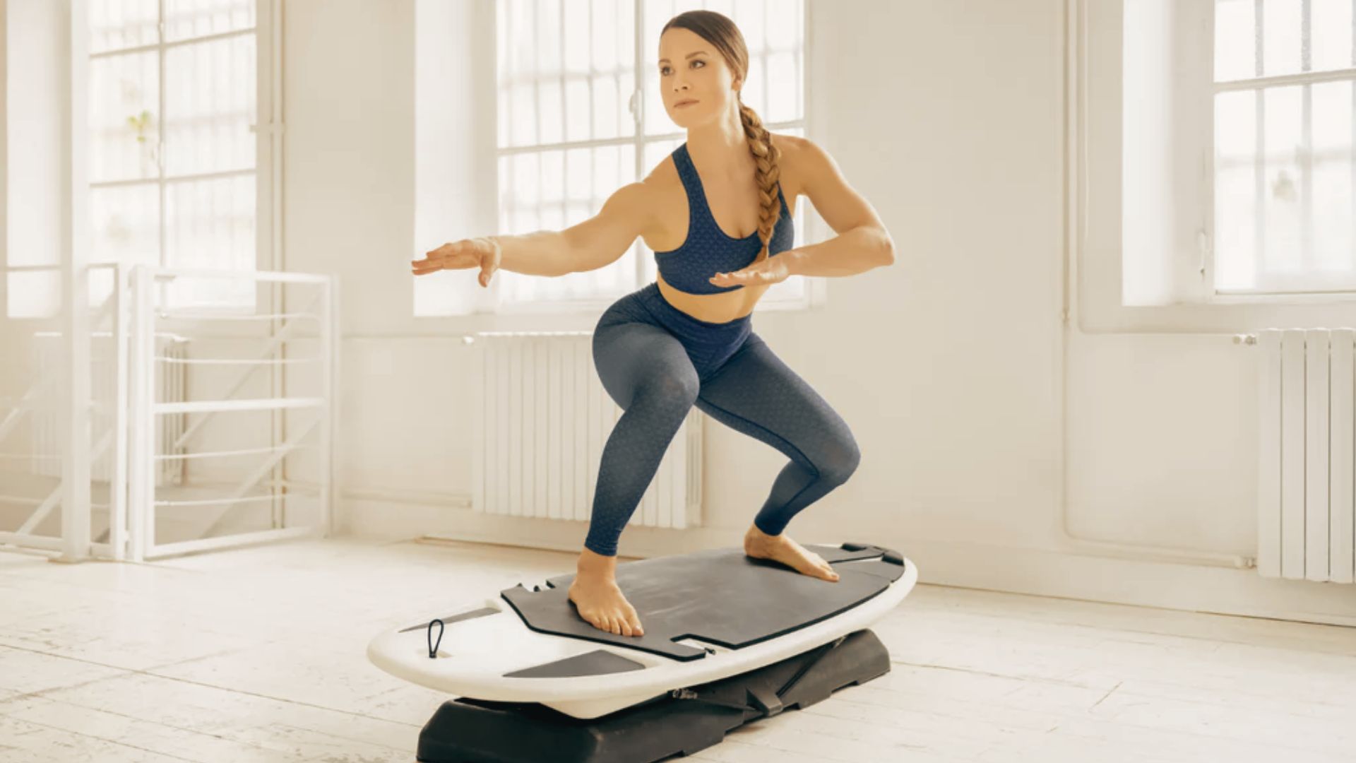 A woman in a bright studio balances in a wide squat stance on a surfboard-style vibration plate.