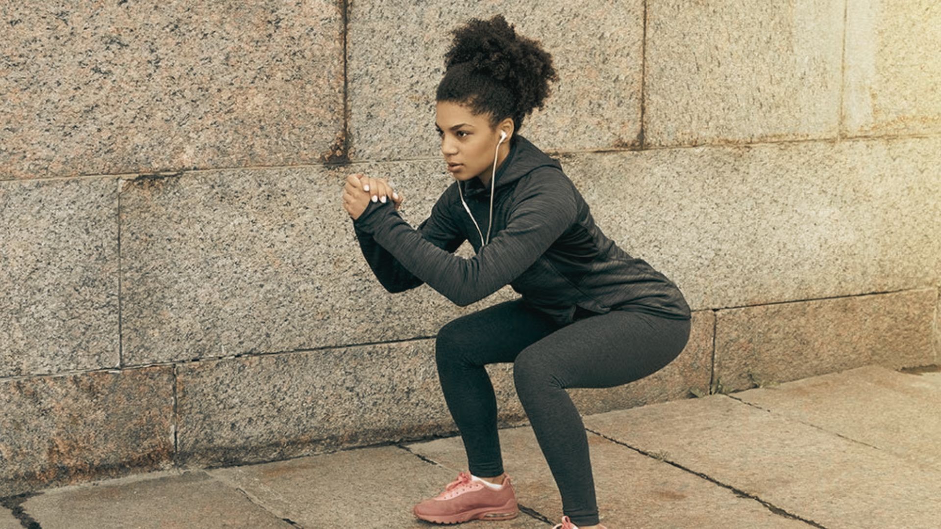 A woman in black athletic wear performs a deep bodyweight squat against a stone wall, demonstrating proper form.