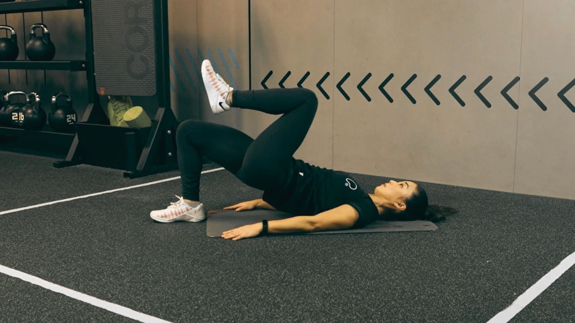 A woman in black leggings lies on a mat performing a single-leg glute bridge in a gym.