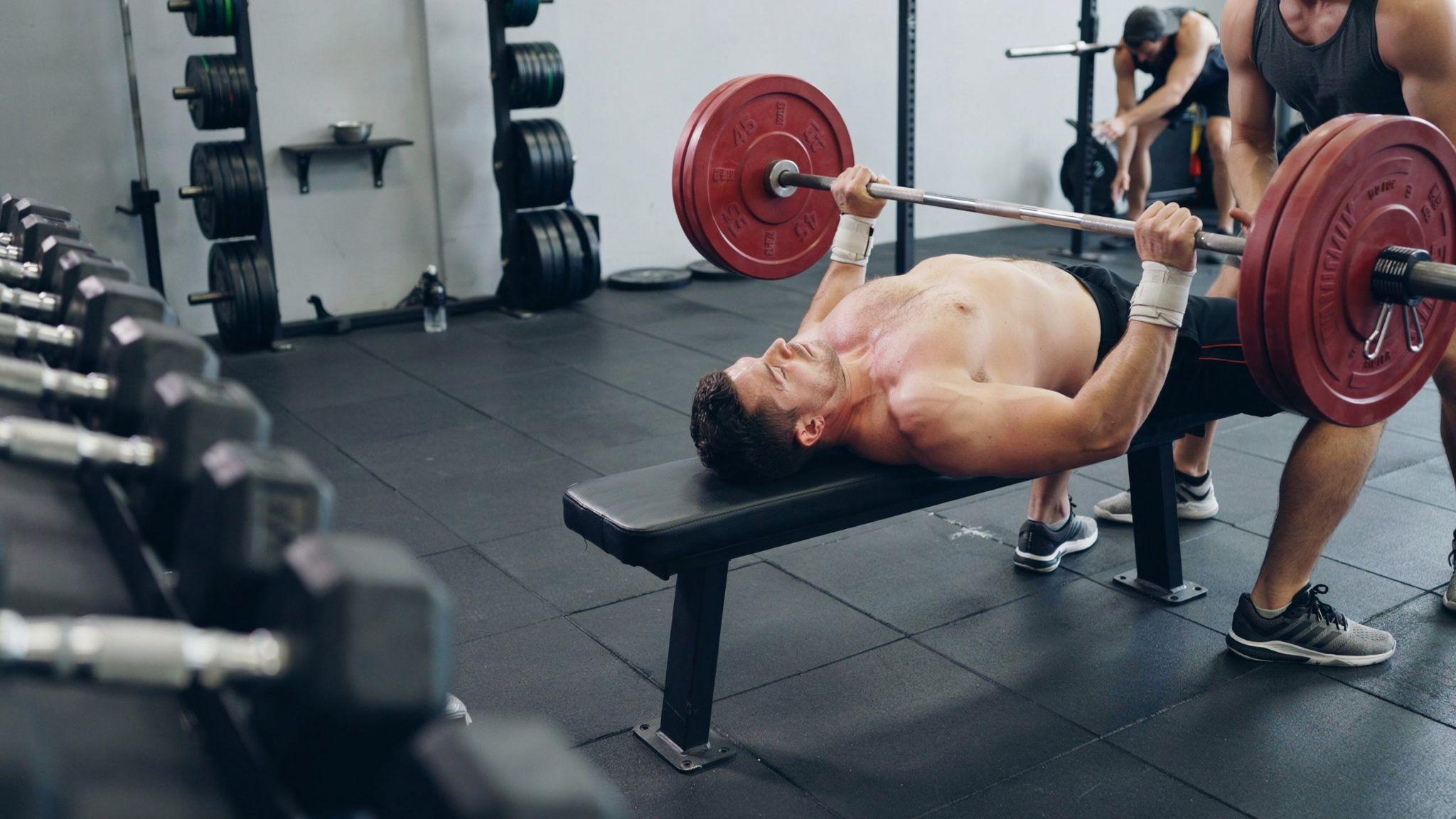 A man performing a barbell bench press on a flat bench in a gym.