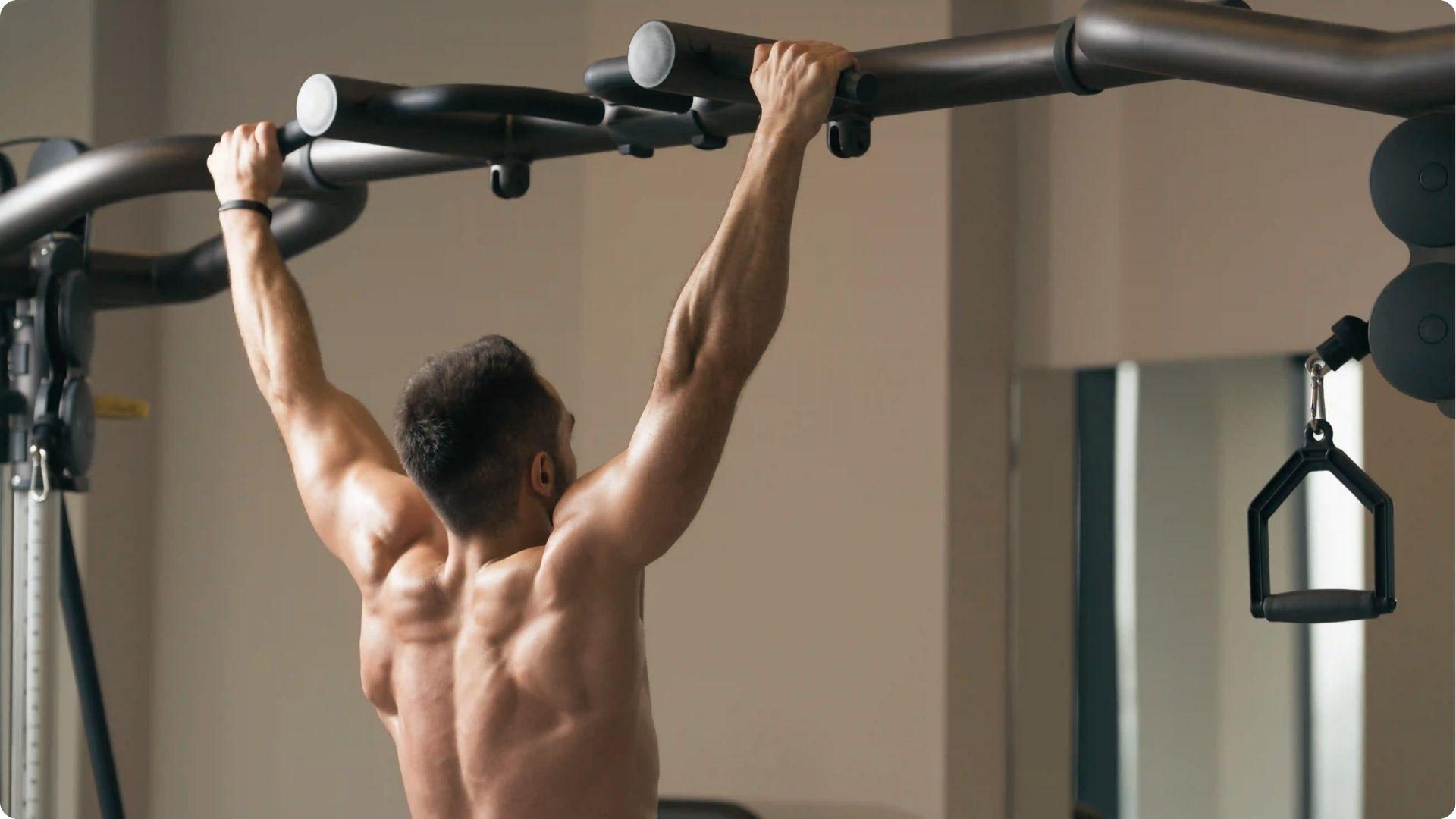 Shirtless man doing wide-grip pull-up on cable machine indoors, back muscles flared