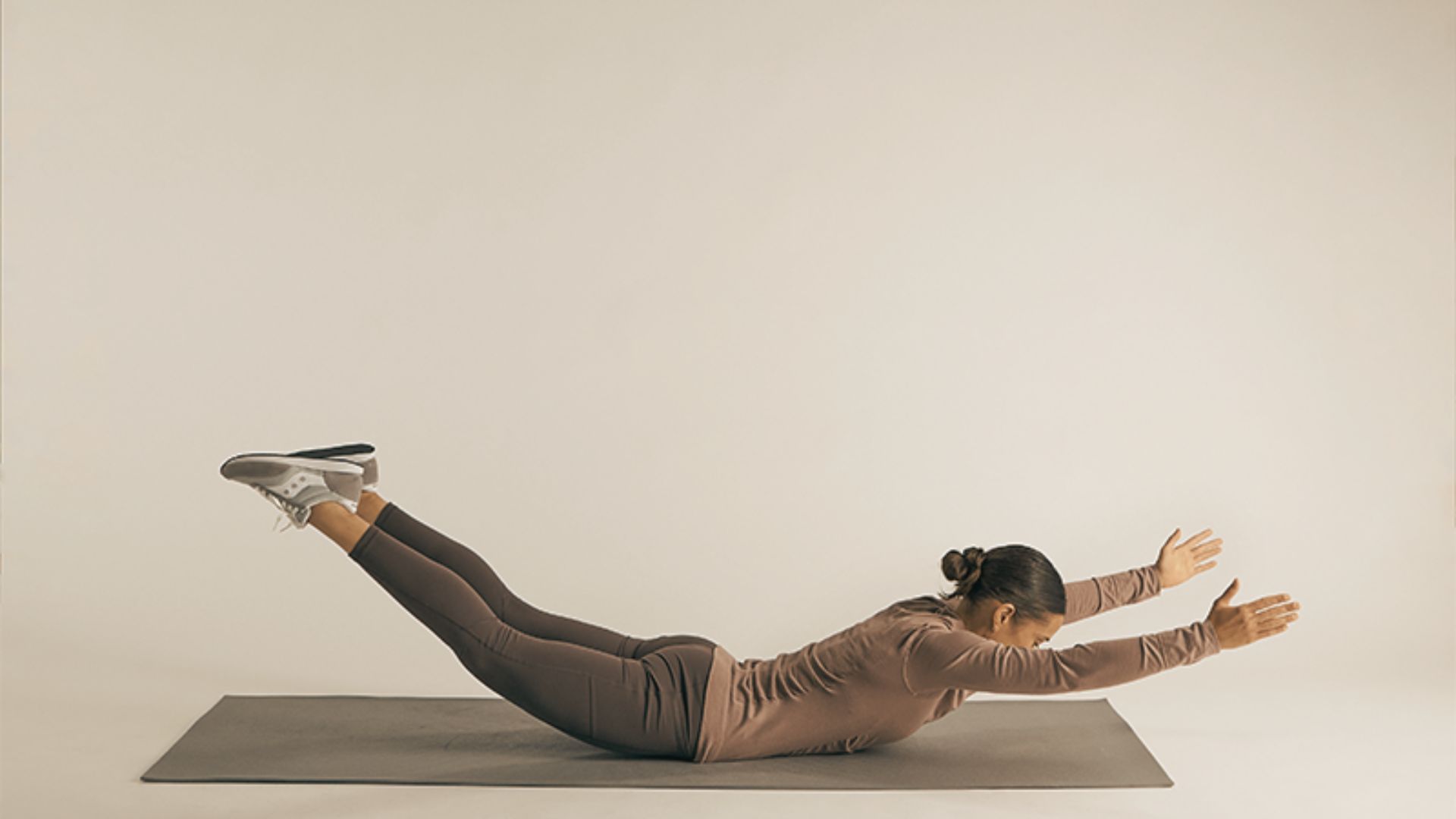 Woman in brown workout set lying face-down on a mat, arms and legs lifted off the ground.