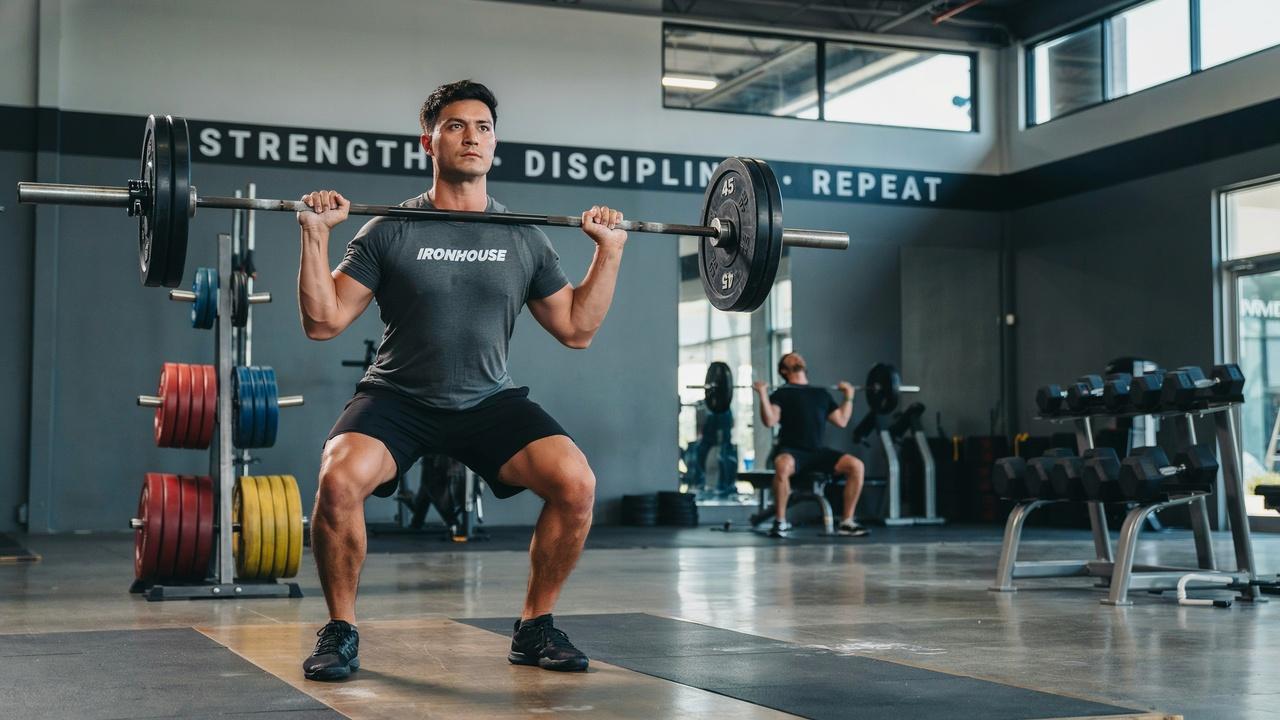 Person doing a barbell squat in a gym with weight racks and dumbbells in the background.
