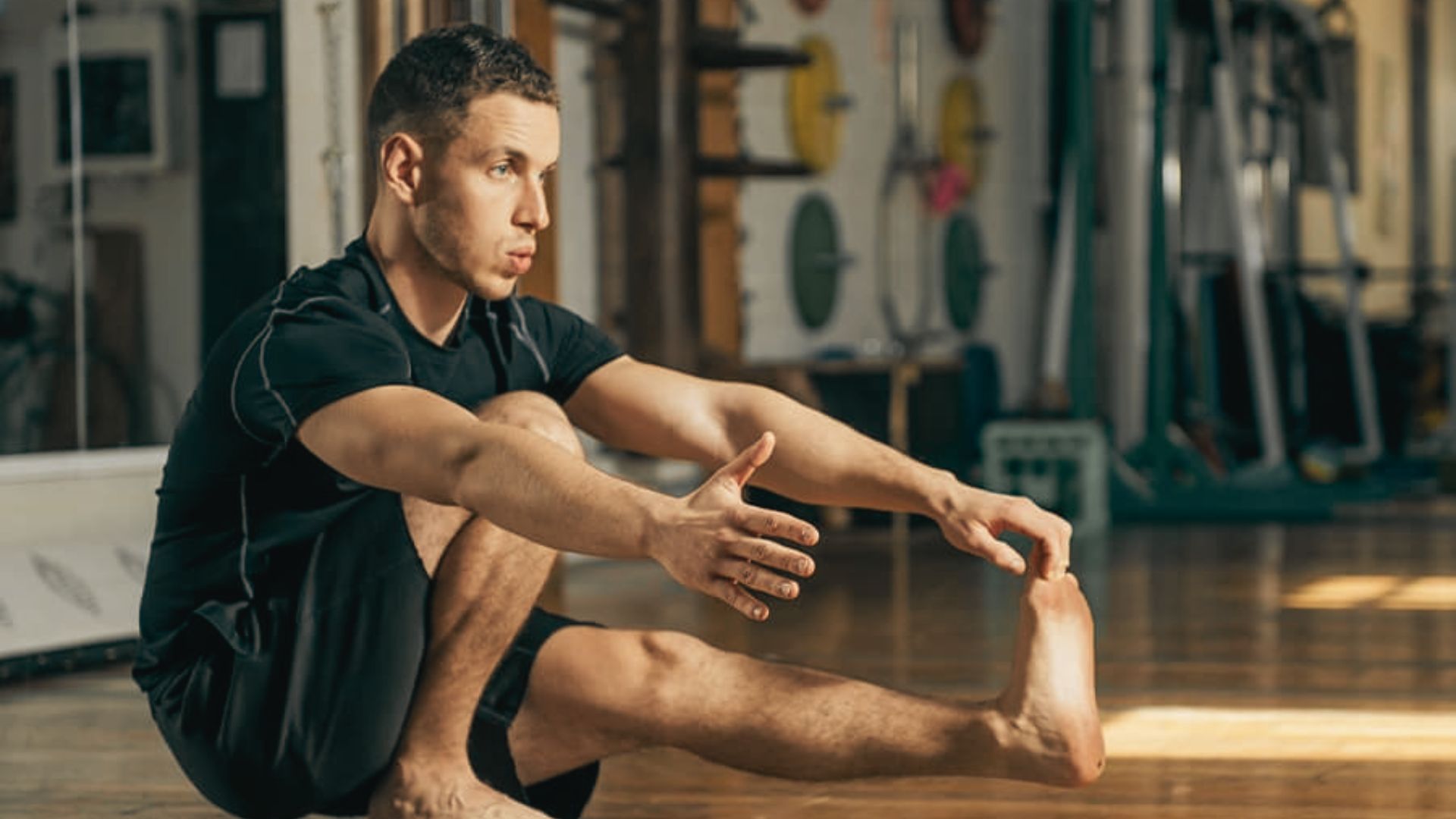 A barefoot man in black workout clothes performs a pistol squat on a gym floor.