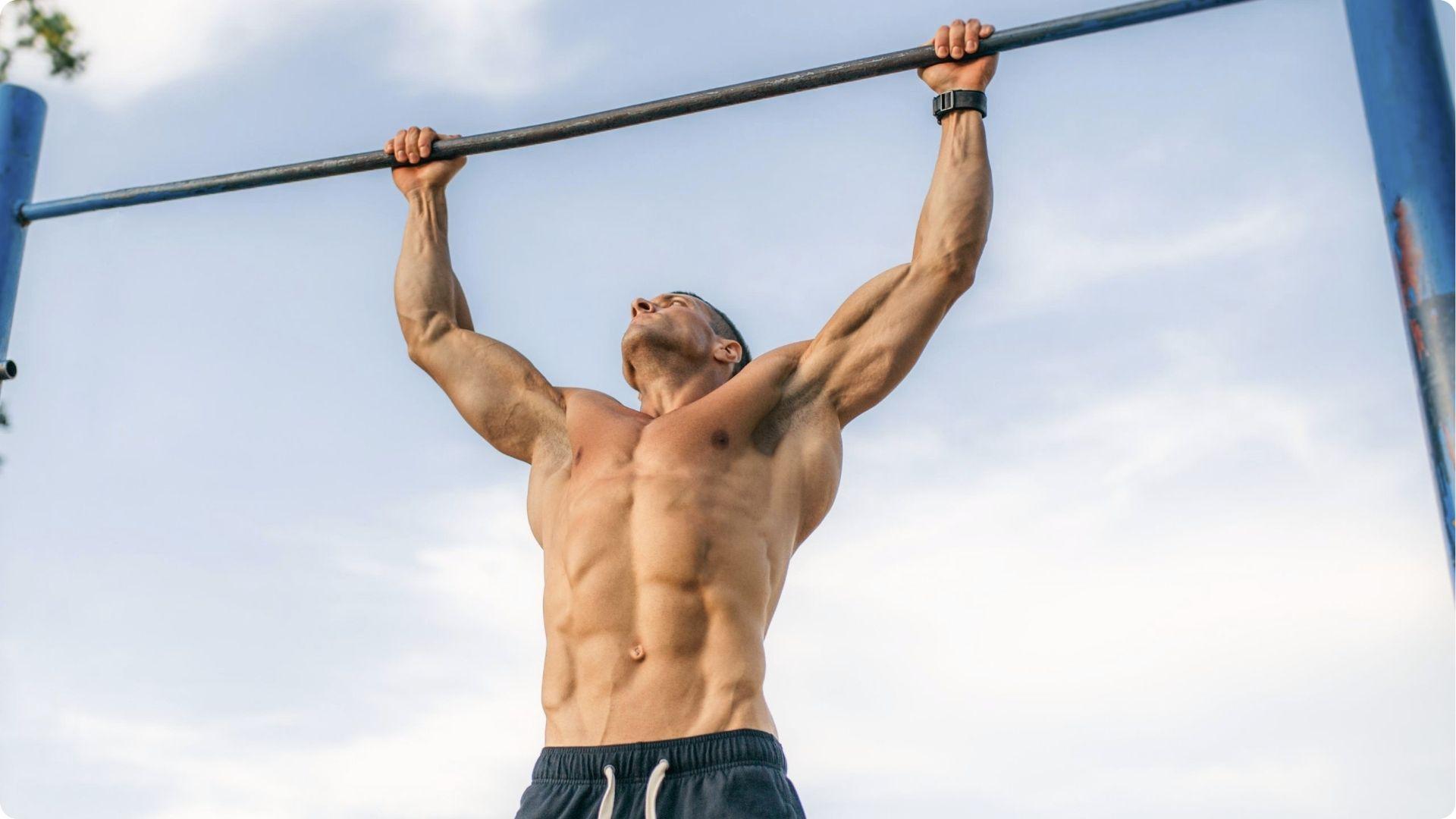 Muscular man performing wide-grip pull-up outdoors, arms and shoulders fully extended