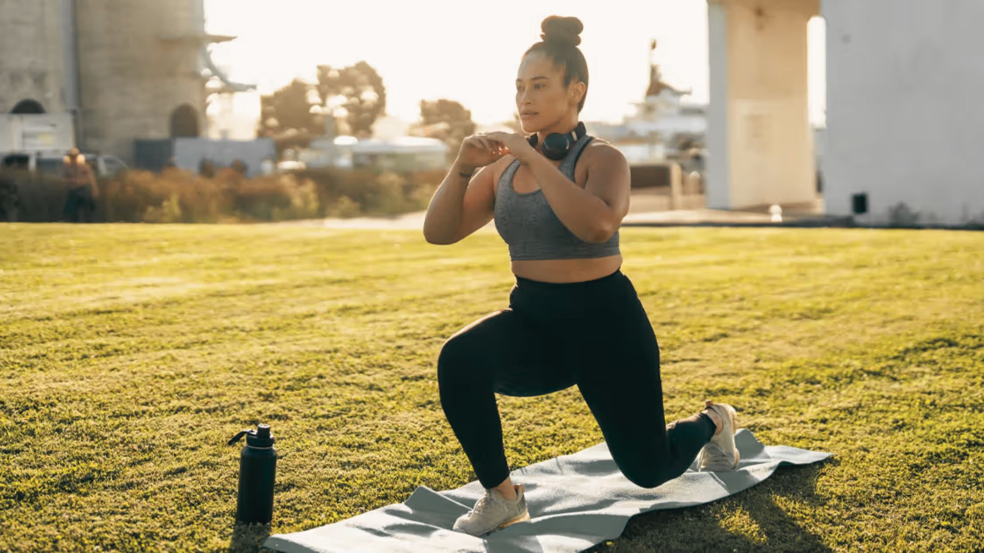 A woman in athletic wear performs a bodyweight lunge on a yoga mat outdoors at sunset.