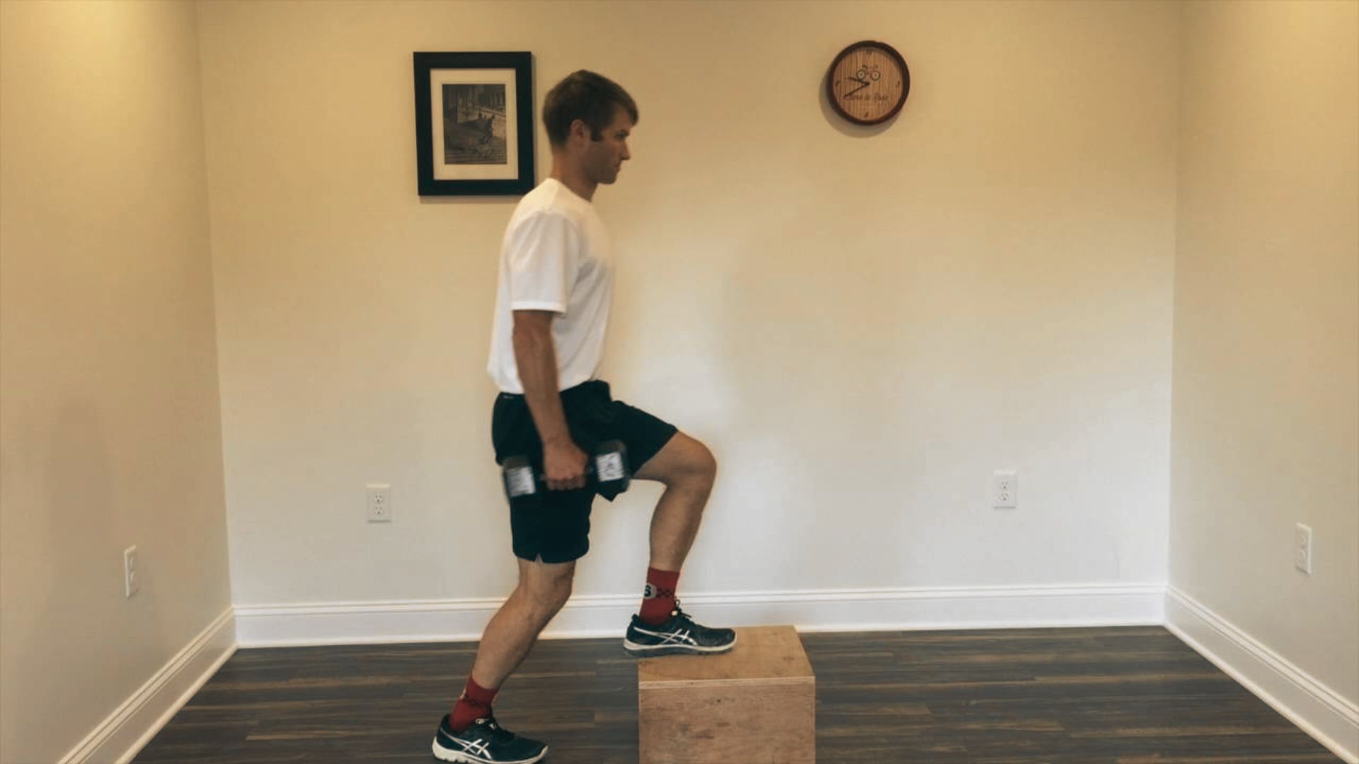 A man in a white shirt holds a dumbbell while stepping onto a wooden box with one leg.