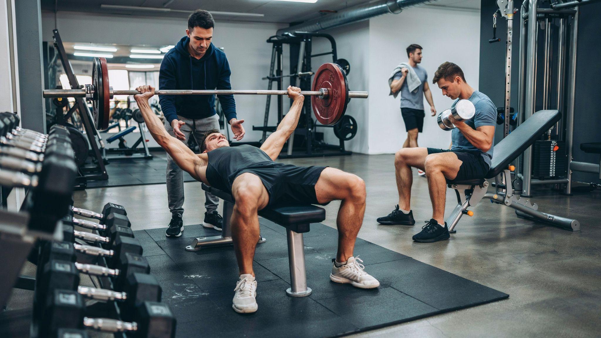 A man performing a barbell bench press with a spotter, while another man lifts dumbbells nearby.