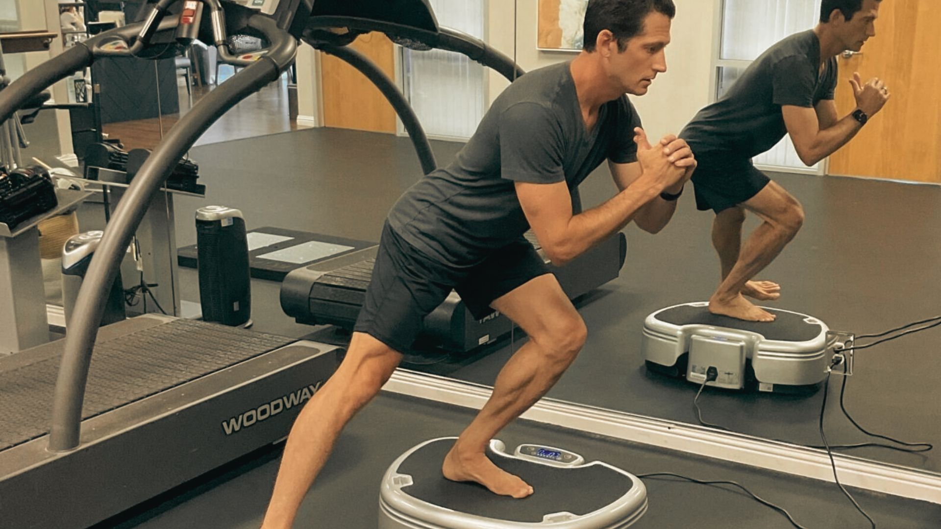 A man in a gym performs a single-leg lunge on a vibration plate, barefoot and leaning forward for balance.
