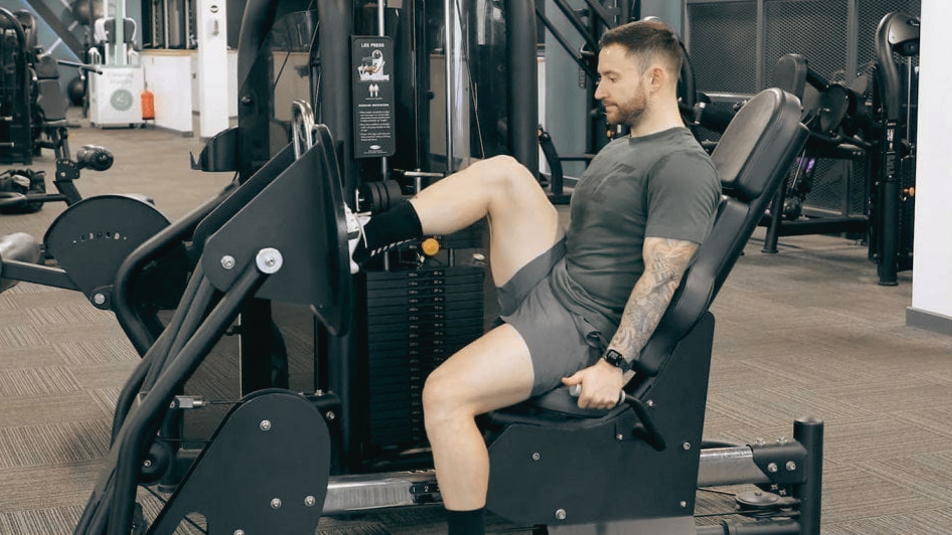 A man in a gray shirt performs a single-leg press on a machine in a gym.