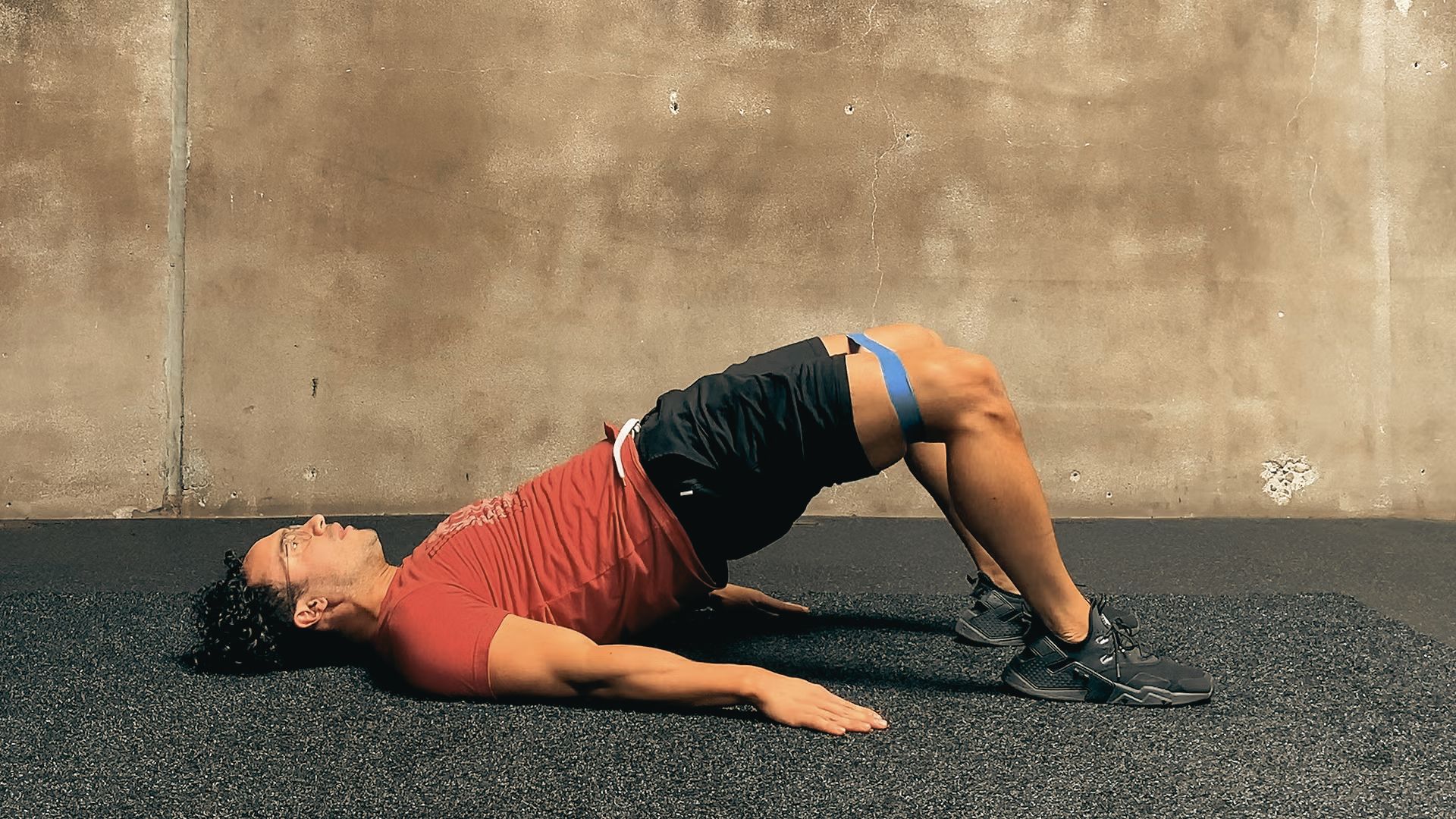 Man in red shirt and black shorts performing a glute bridge with a blue band above his knees.