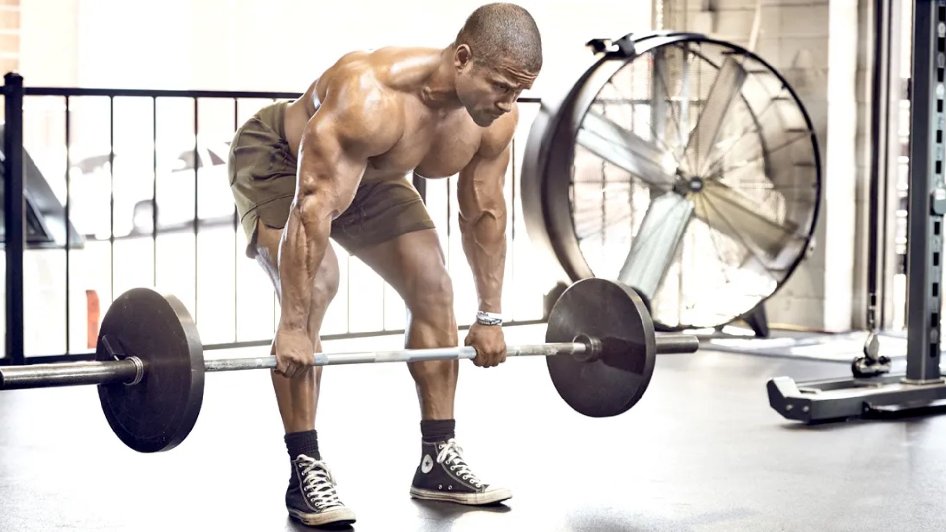 Muscular man in shorts executing a barbell deadlift in a gym with a large fan in the background.