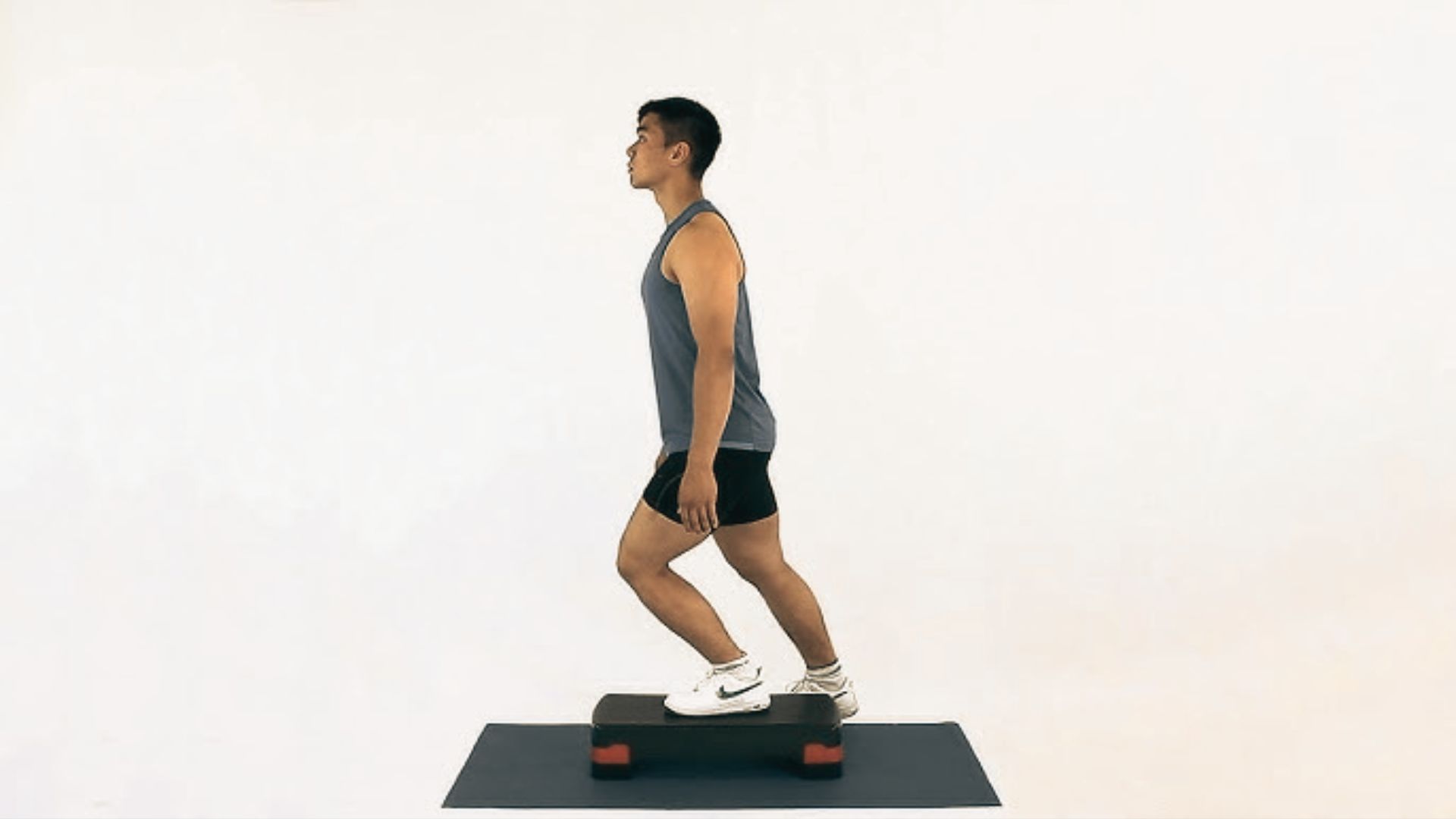 A man in a gray tank top steps up onto a platform with one leg against a white background.